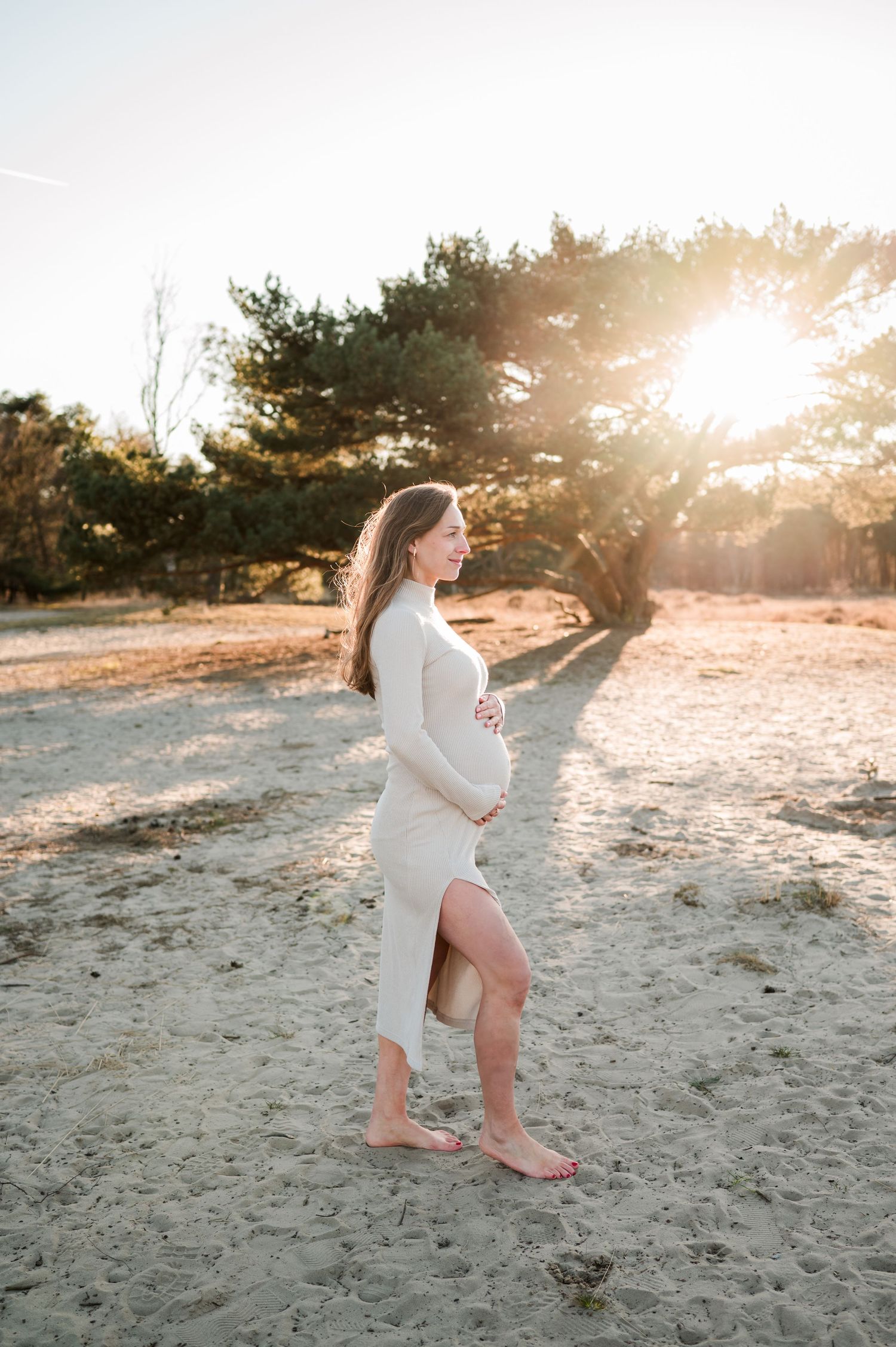 A pregnant woman in a white dress stands on a sandy beach during golden hour sunset with sunlight filtering through trees.