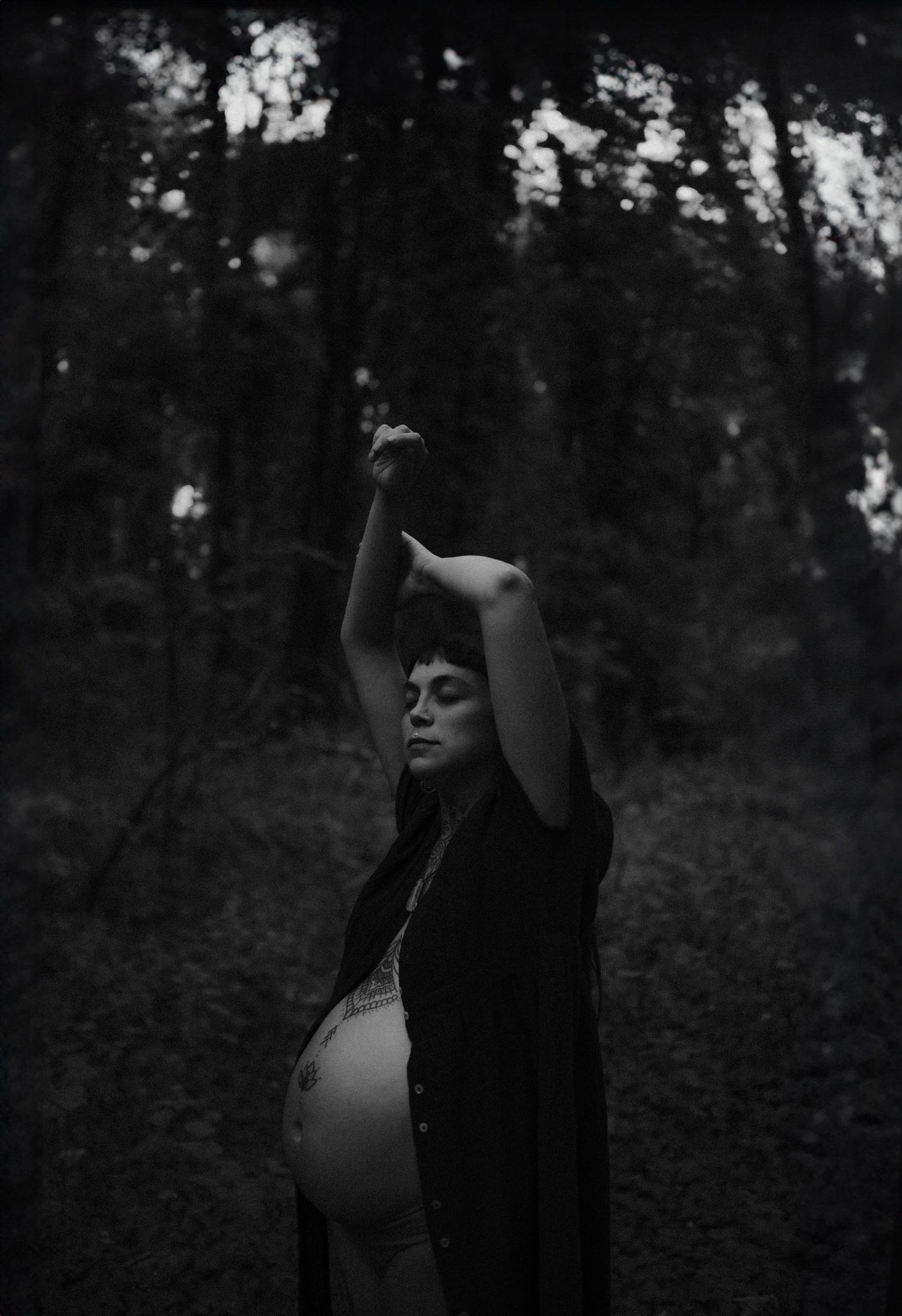 Dramatic black and white maternity portrait with figure reaching skyward in forest.