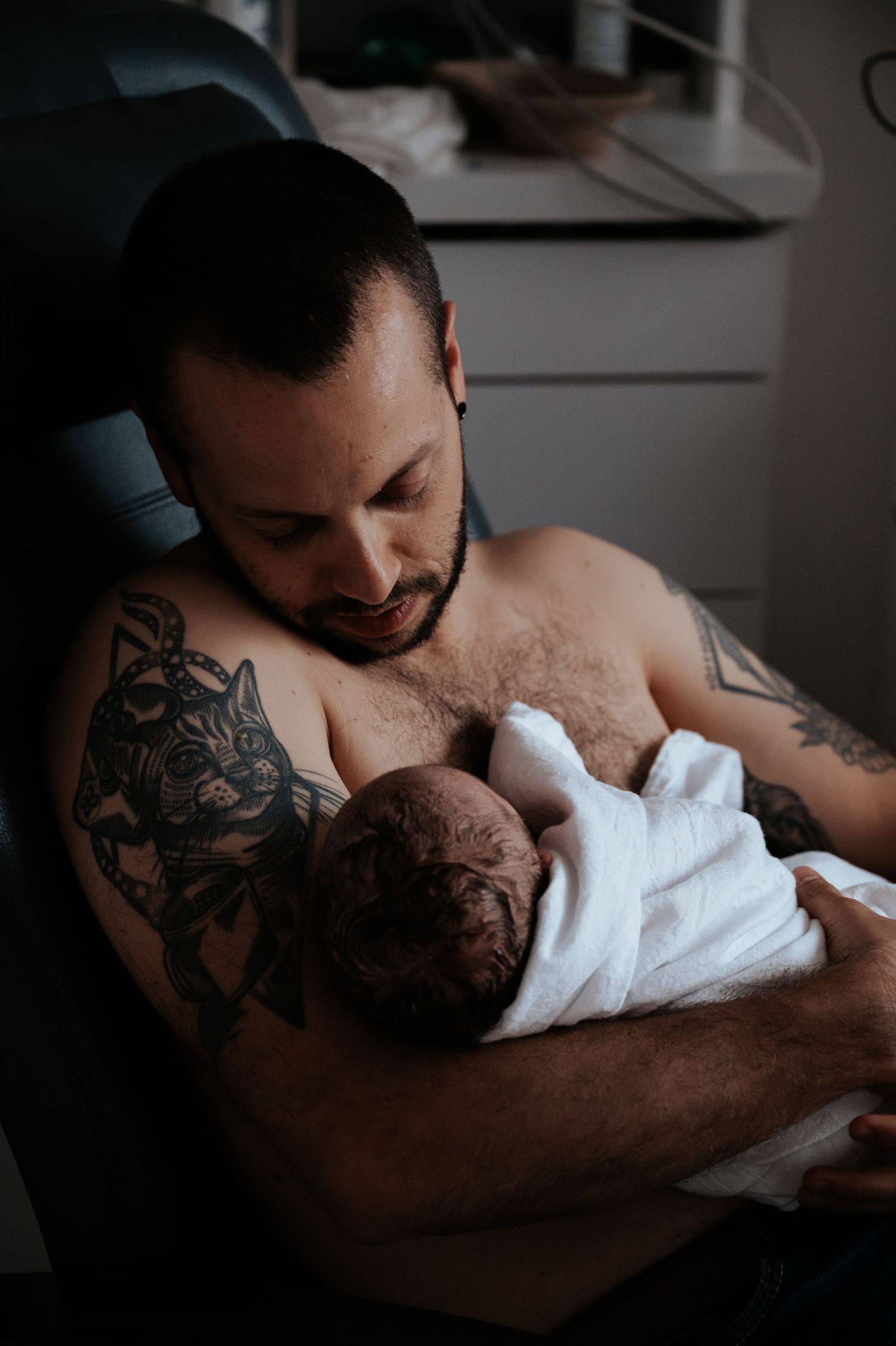 A person with chest tattoos tenderly holds a newborn baby wrapped in a white blanket in an intimate indoor setting.