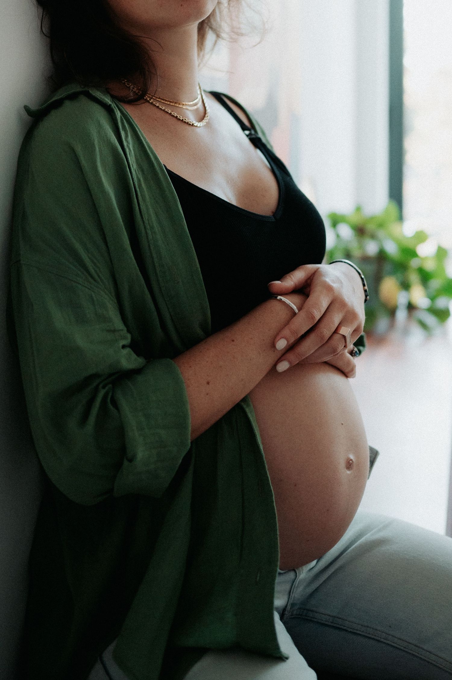Artistic maternity photo showing a pregnant belly with woman wearing green cardigan and black top by window.