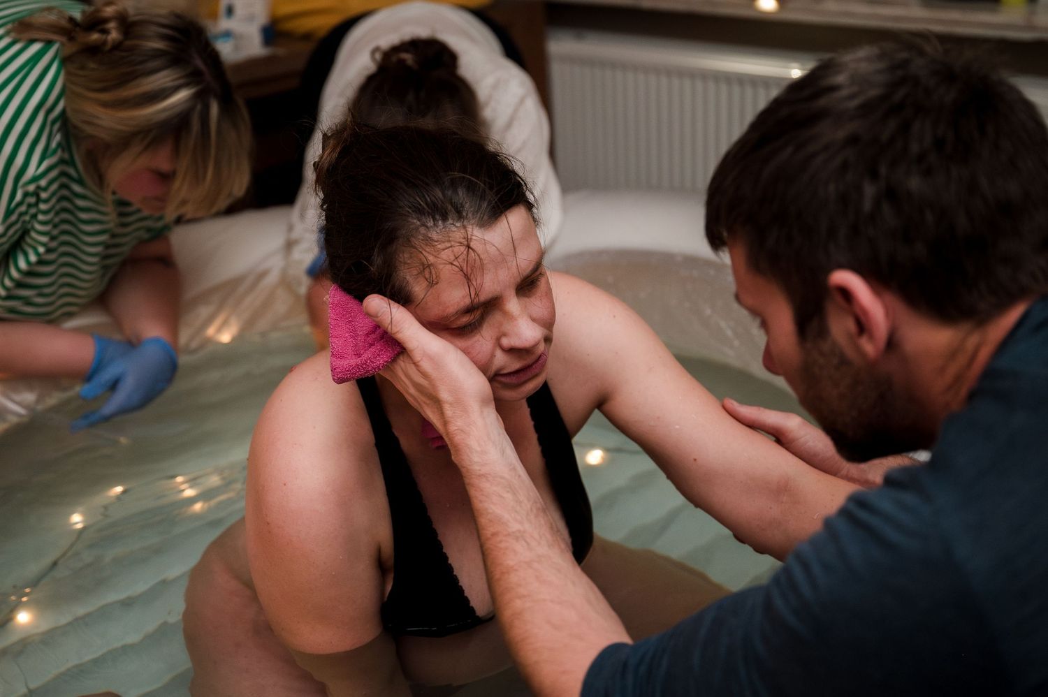 Woman in birthing pool supported by partner and midwife during water birth labor.