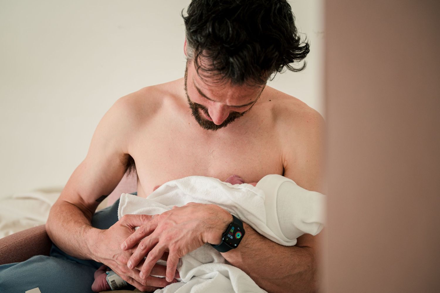 A shirtless adult gently embraces a white blanket bundle while sitting in soft lighting.