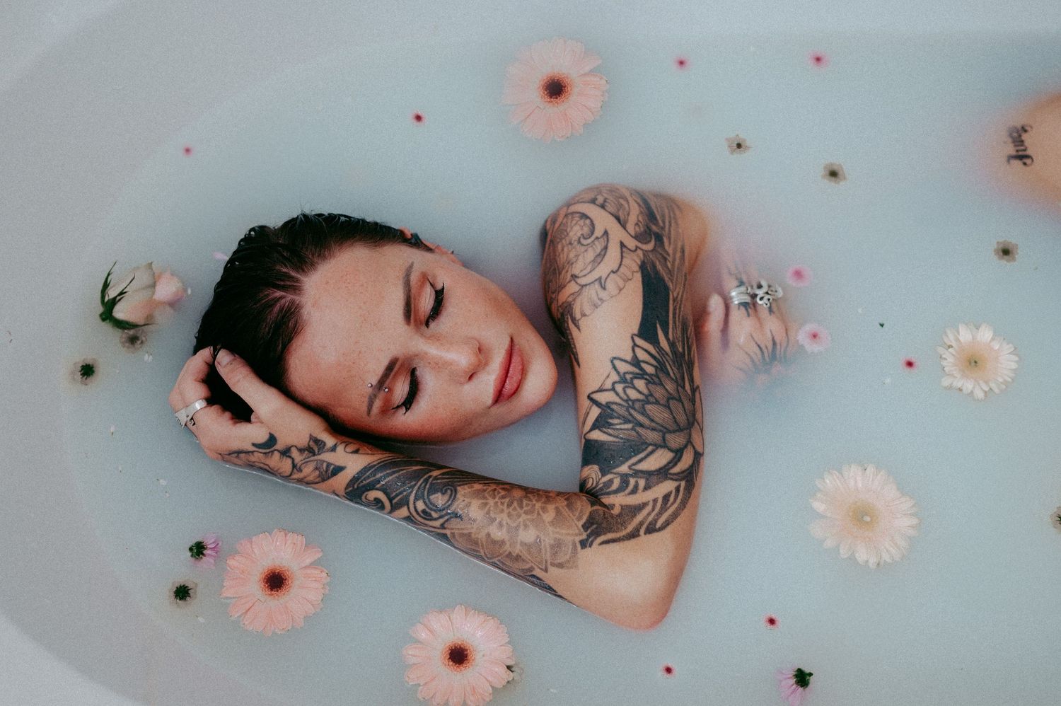 Artistic bathtub portrait with pink flowers floating in milky water and visible tattoo sleeve.