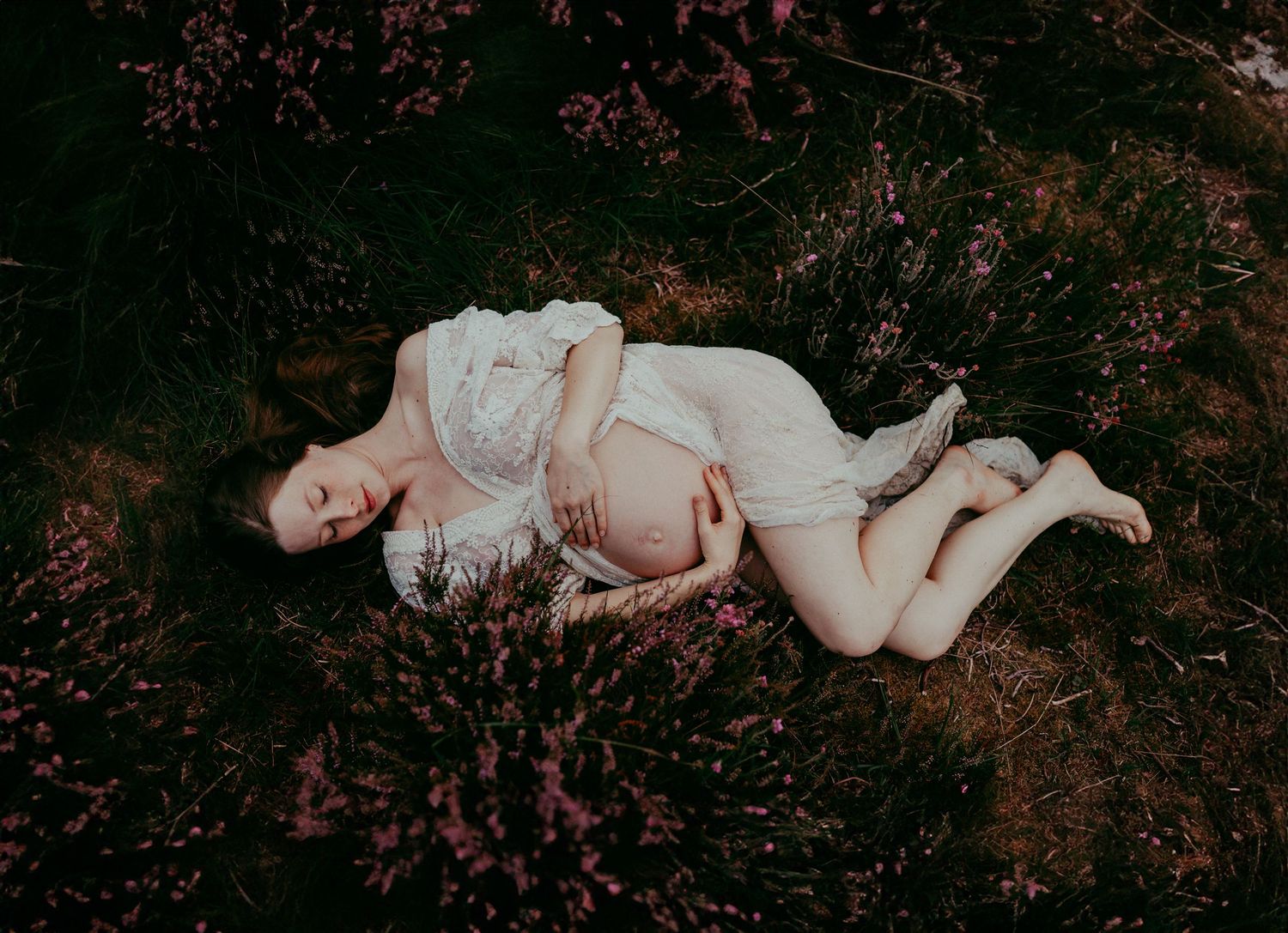 Artistic maternity portrait with subject lying among spring blossoms wearing a white lace dress.