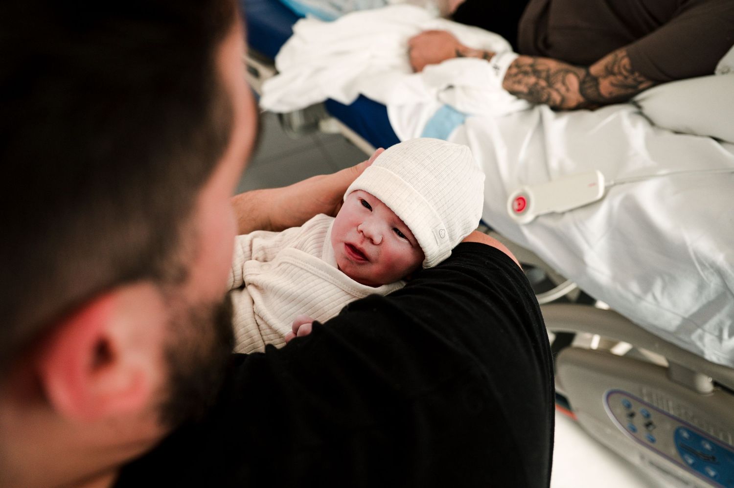A newborn baby swaddled in white cloth lies in a caregiver's arms while being held in a medical setting.