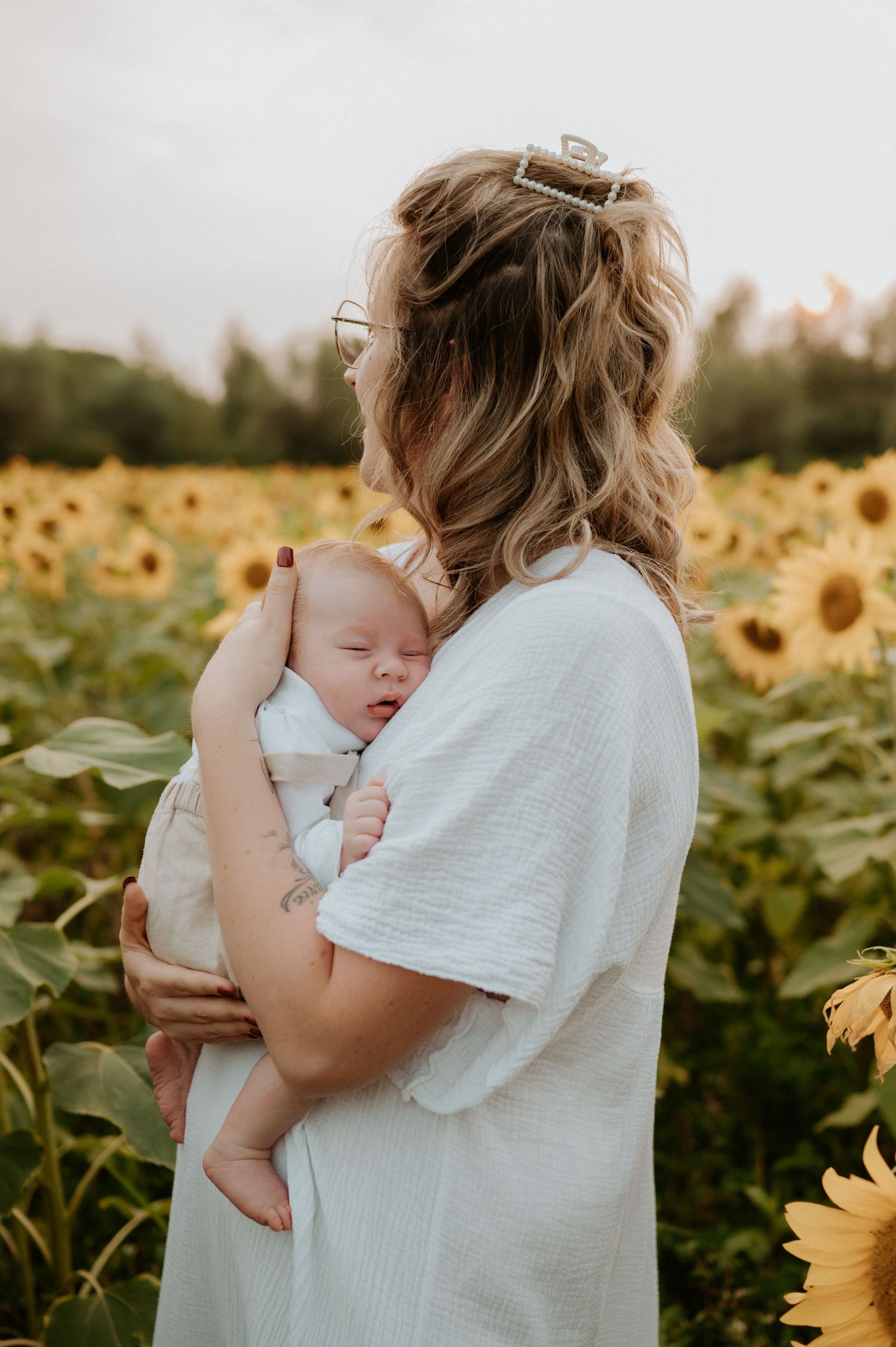 Mother holding sleeping baby in sunflower field wearing white shirt.