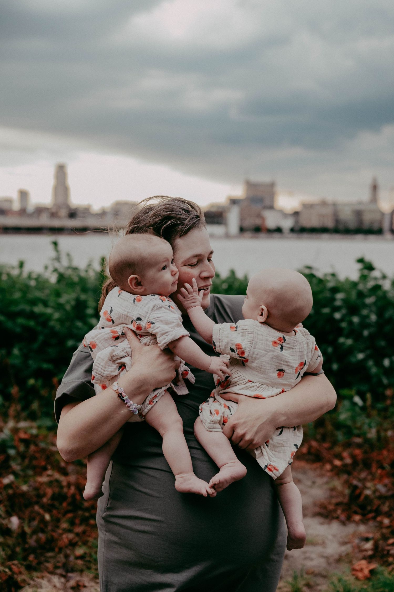 Parent holding twin babies in floral outfits against urban waterfront backdrop.
