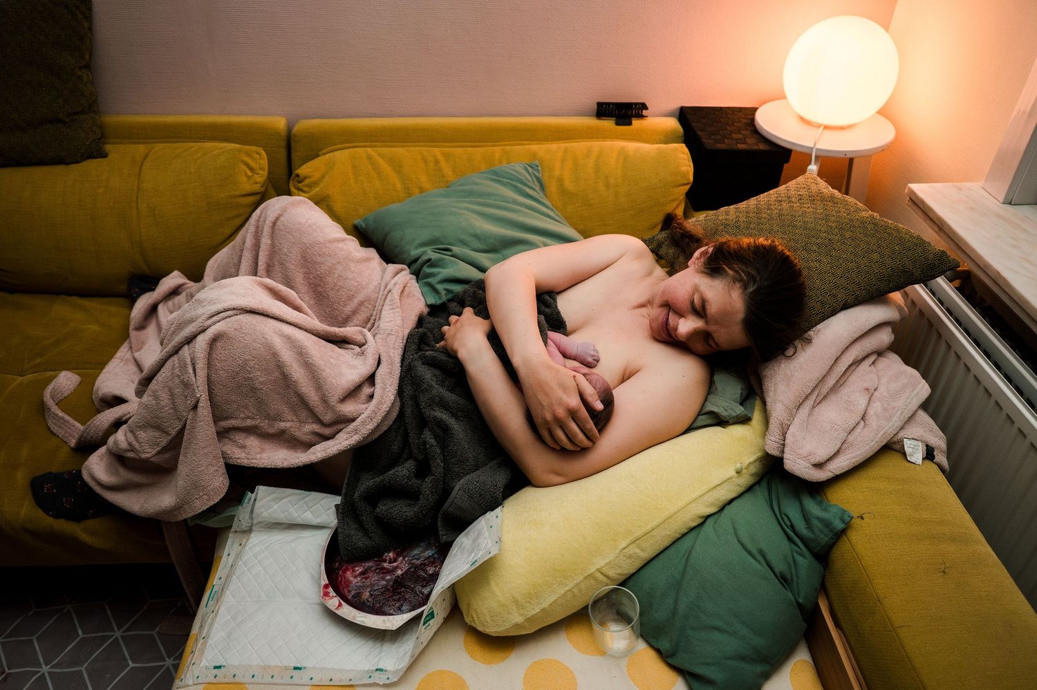 Person resting on a yellow sofa surrounded by colorful pillows and blankets in a cozy living room with warm lighting.