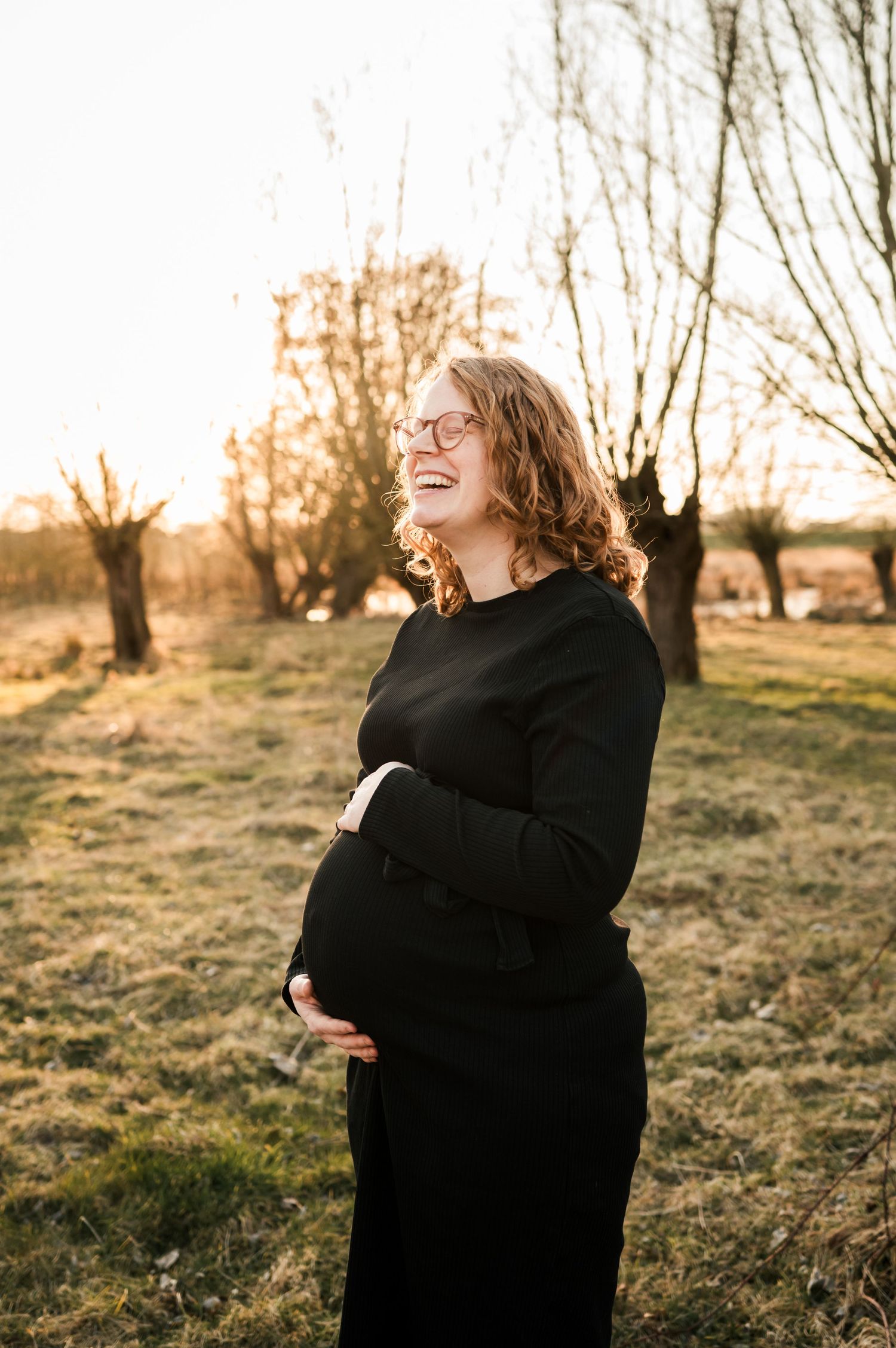 Joyful pregnant person laughs in golden sunlight wearing black sweater dress outdoors at sunset.