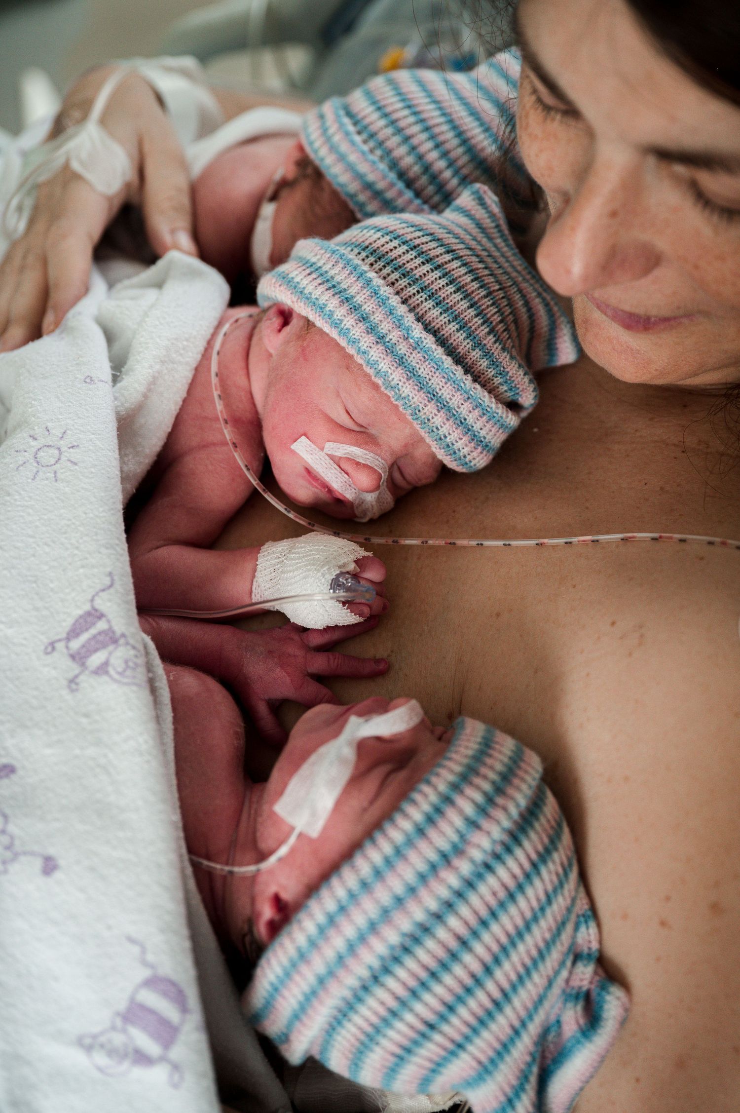 Three newborn babies wearing matching blue and white striped knit caps rest against their parent after birth.