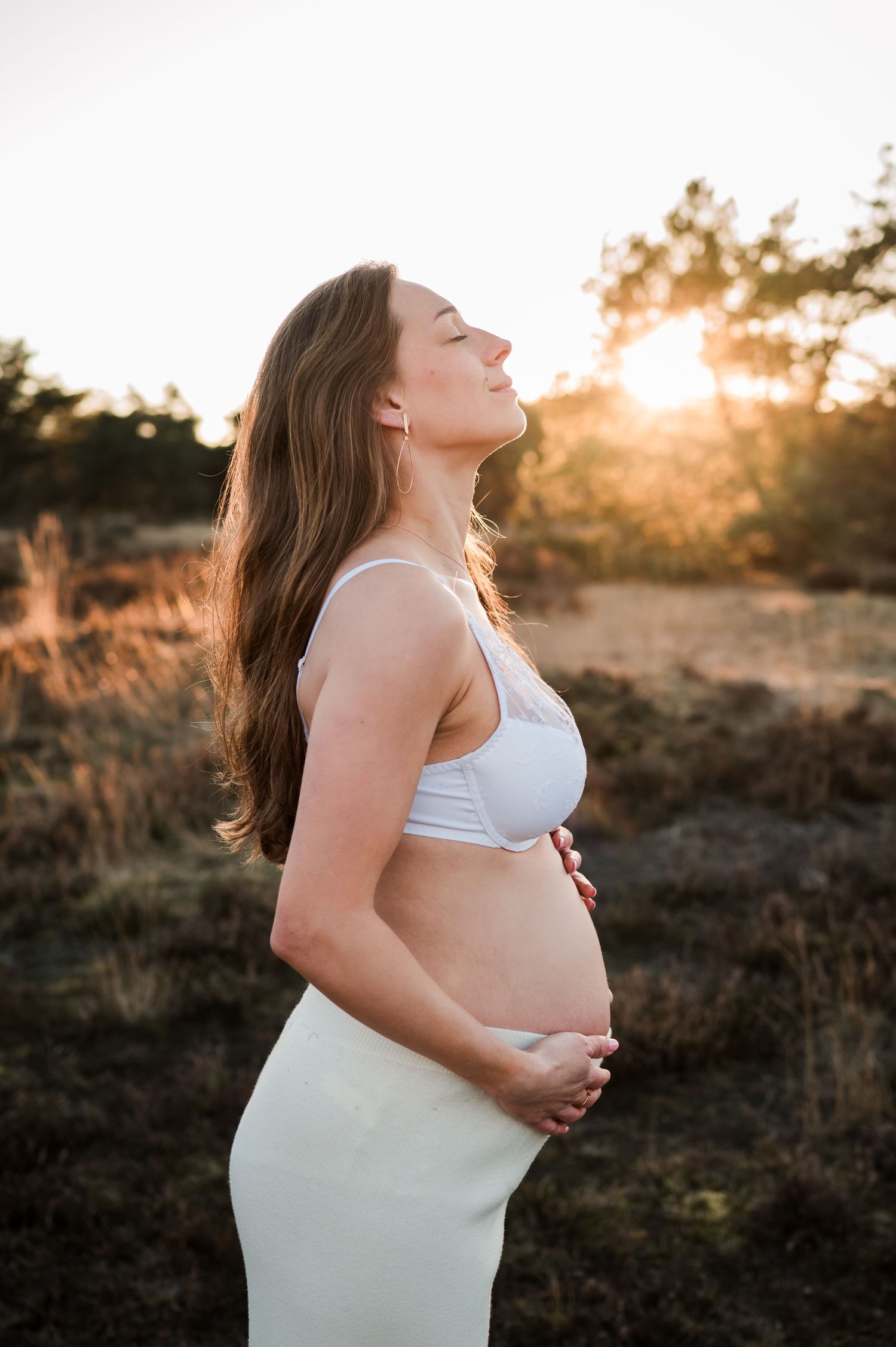 A pregnant woman in white attire poses serenely against a golden sunset in a natural outdoor setting.