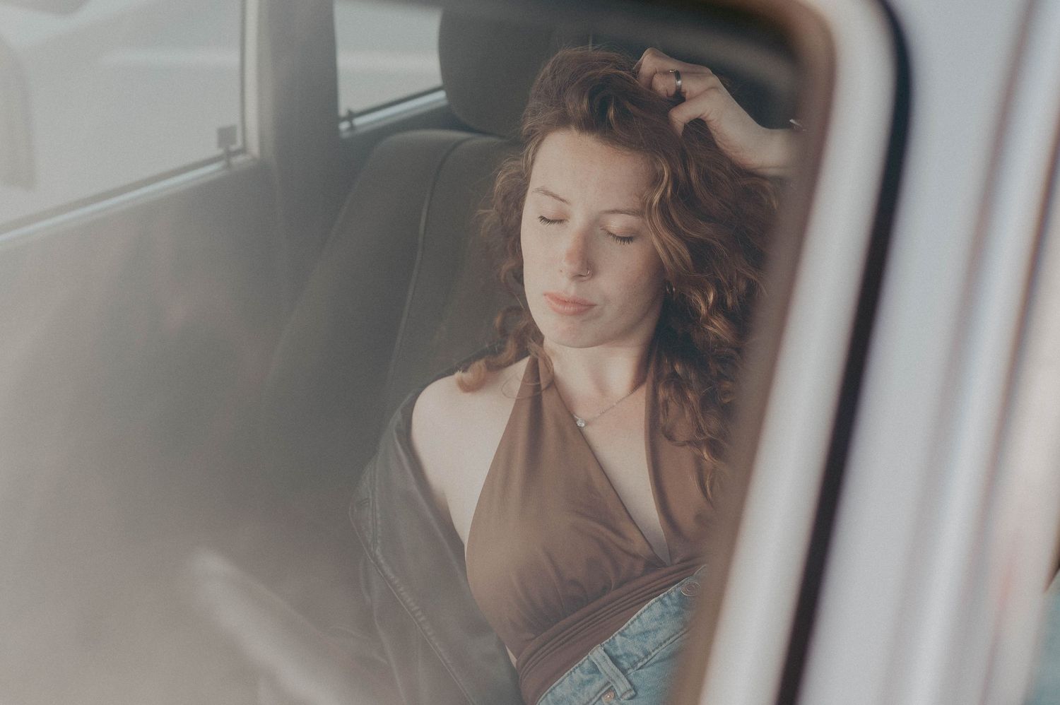 A person with wavy brown hair sits thoughtfully in a car, viewed through a window with soft diffused lighting.