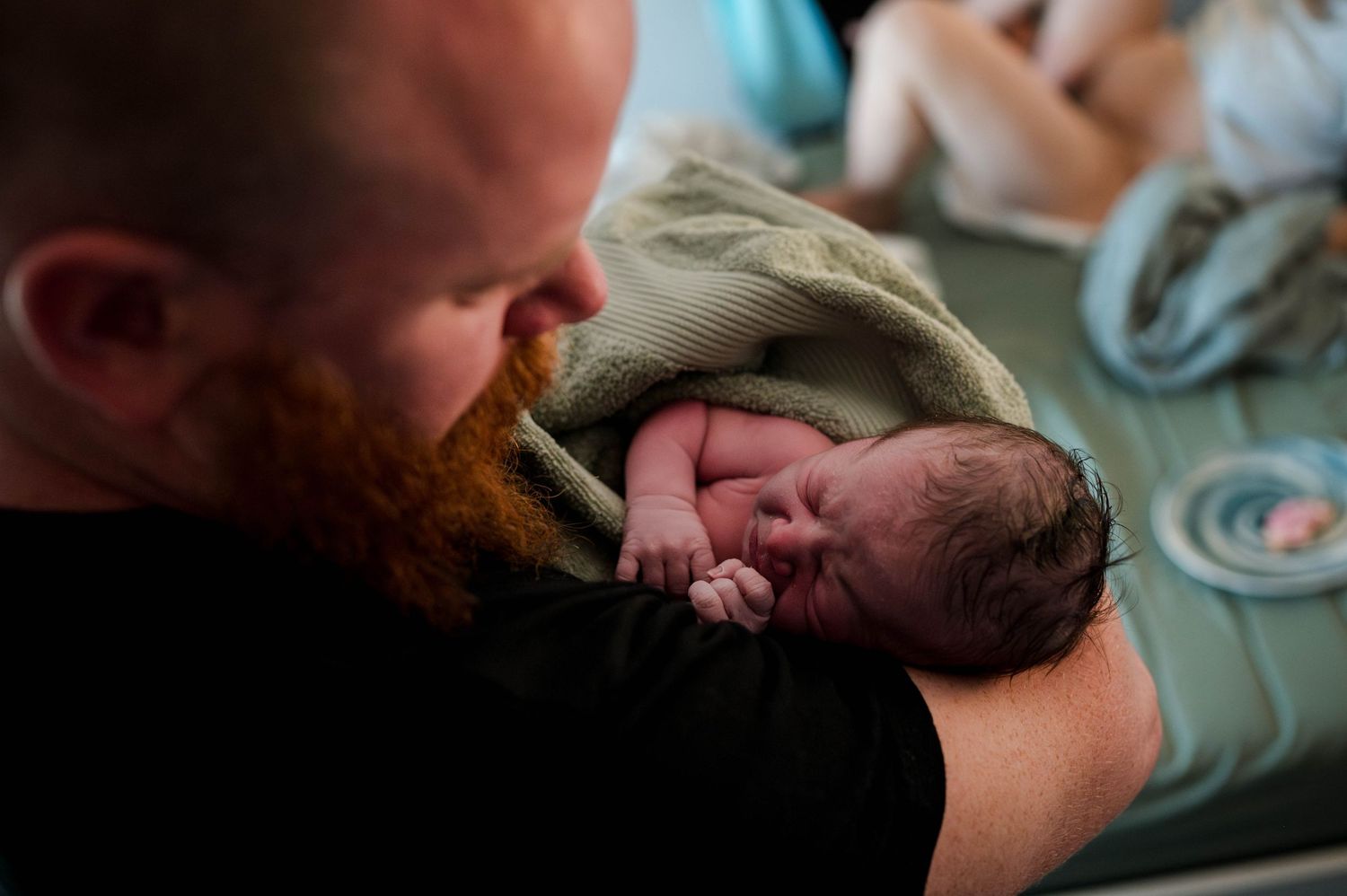 A newborn baby wrapped in a beige blanket rests in someone's tattooed arms during an intimate moment.