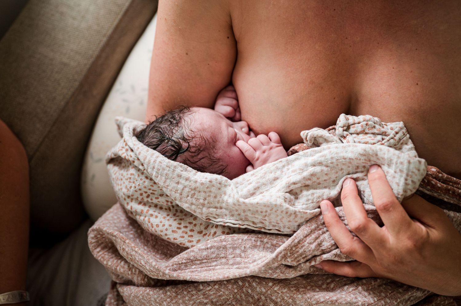 A newborn baby wearing a knitted blanket lies peacefully on a soft bed in warm natural lighting.