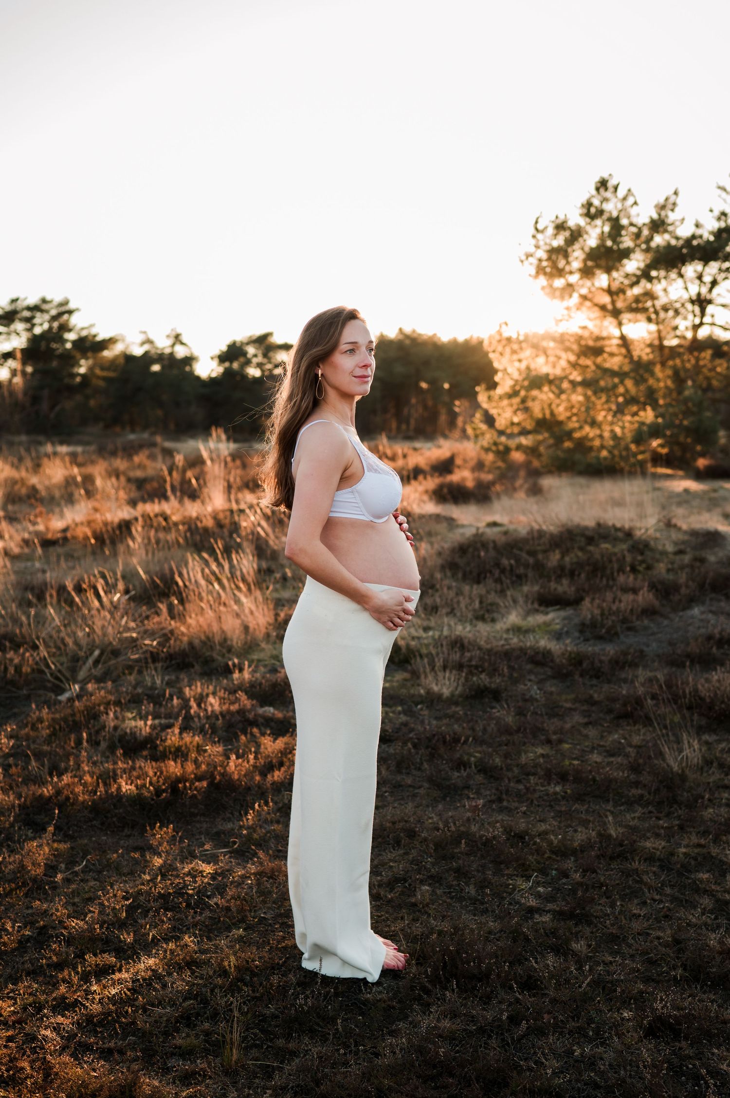 Expectant mother in white dress poses during golden sunset in field with tall grass and pine trees in background.
