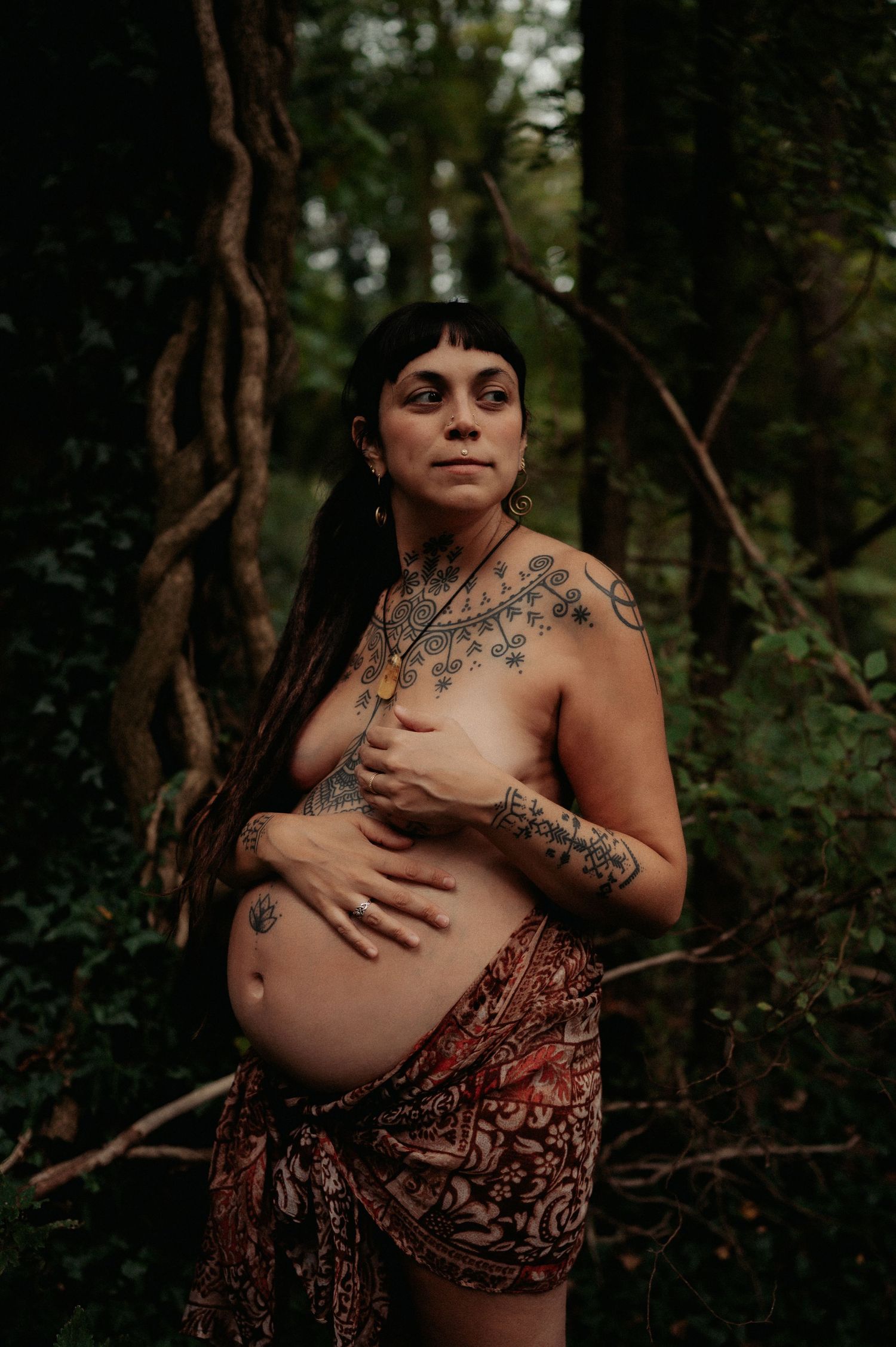 Artistic maternity portrait in forest showing pregnant figure with tattoos in patterned wrap.