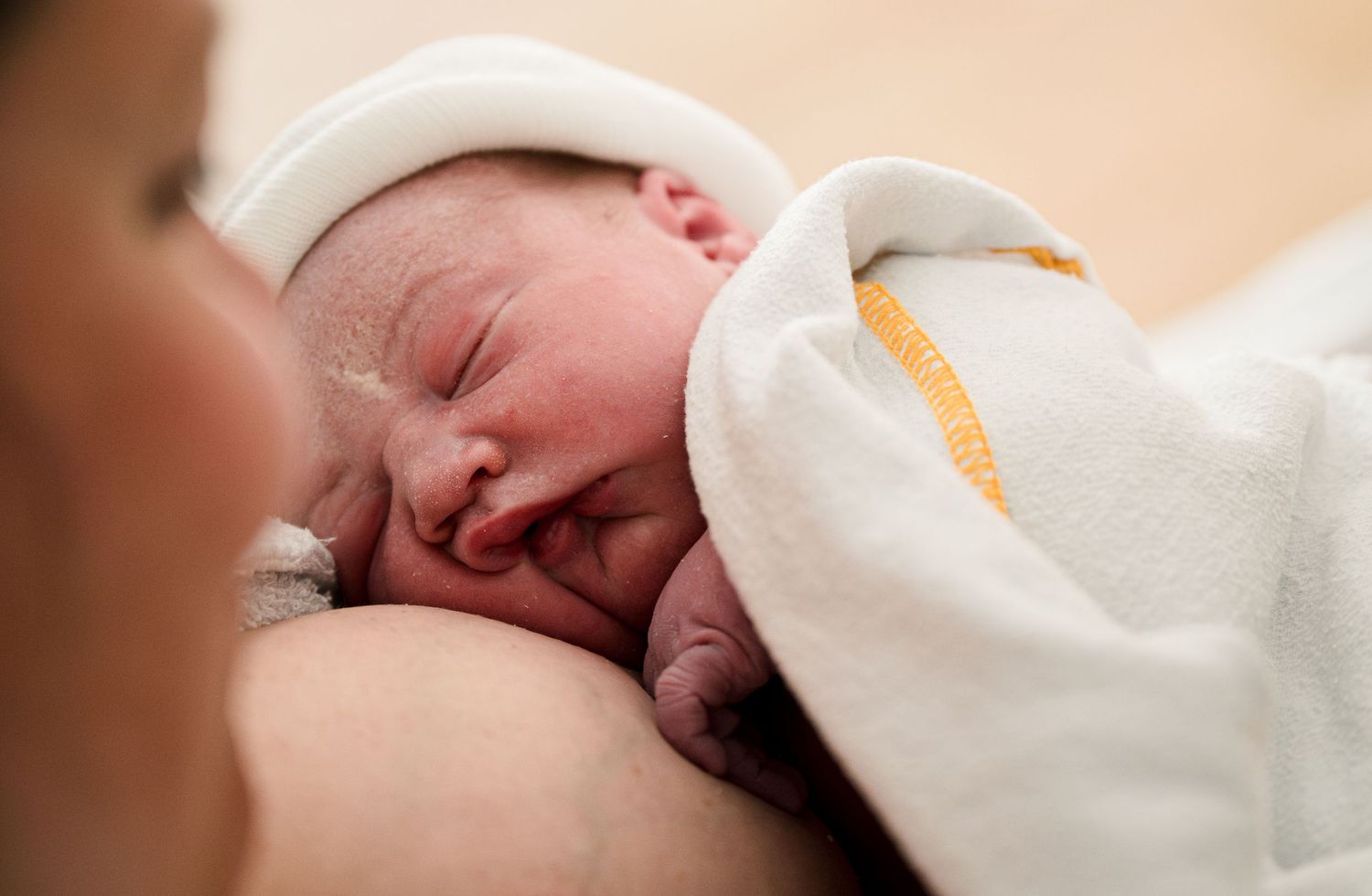 An adorable sleeping newborn baby in white swaddling cloth is resting peacefully against a warm beige background.