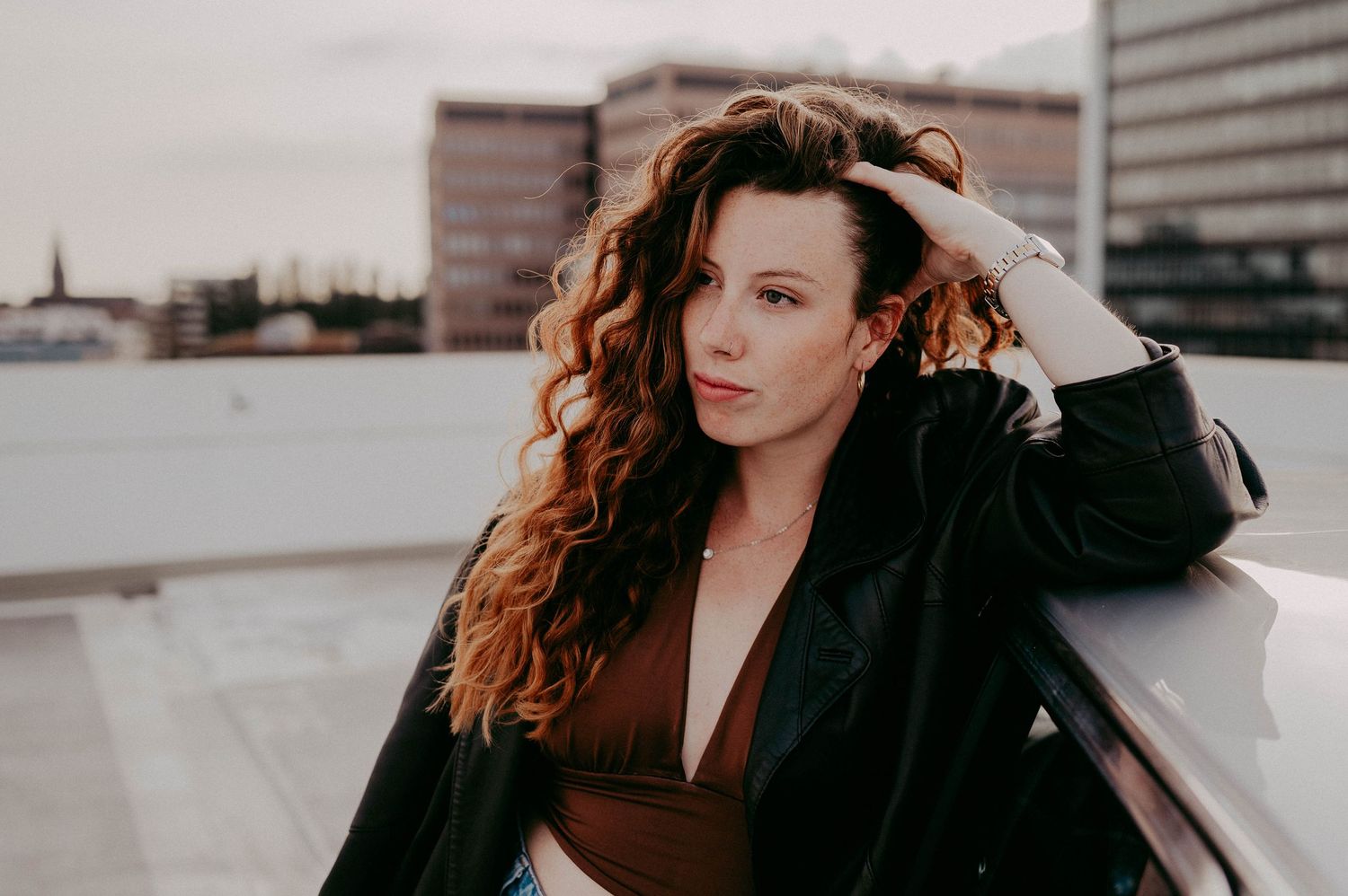 Fashion portrait featuring long curly hair and black coat against an urban rooftop setting.