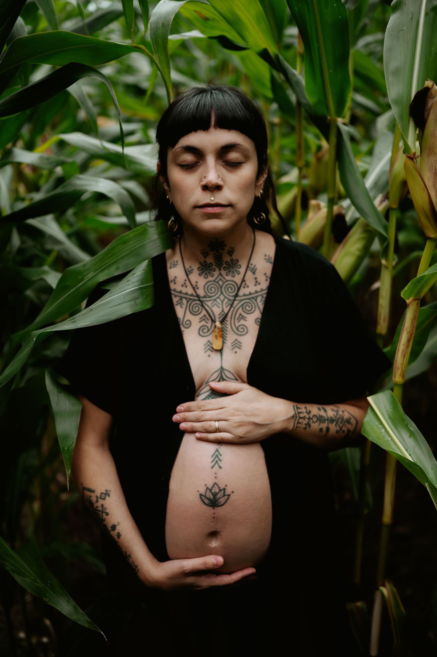 Portrait of a pregnant person wearing a black top with tattoos, standing amid lush green foliage in a garden setting.