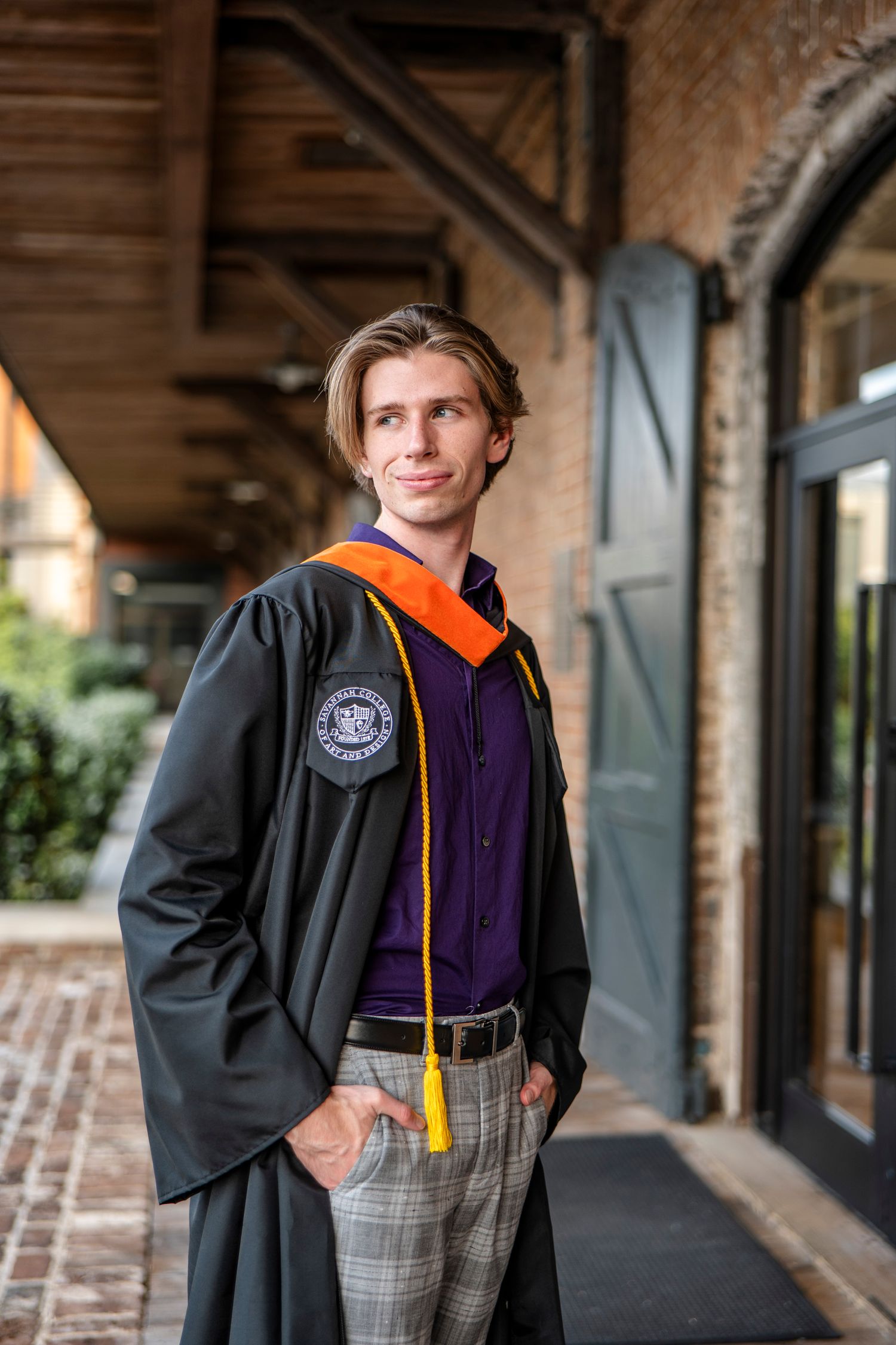 A graduate poses in academic regalia with orange and yellow stoles in front of a brick building.