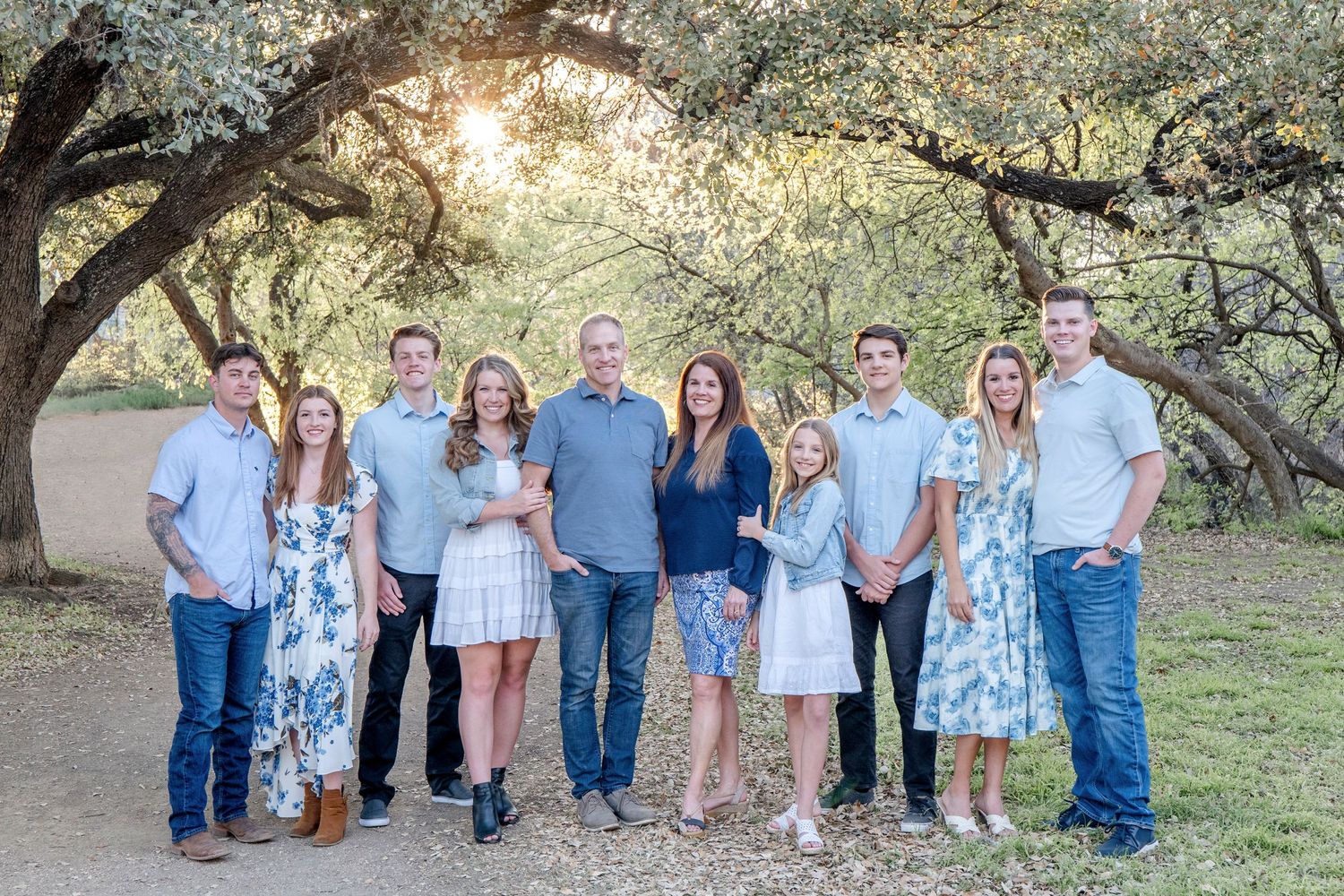 A group in coordinated blue and white outfits poses together outdoors under oak trees at sunset.