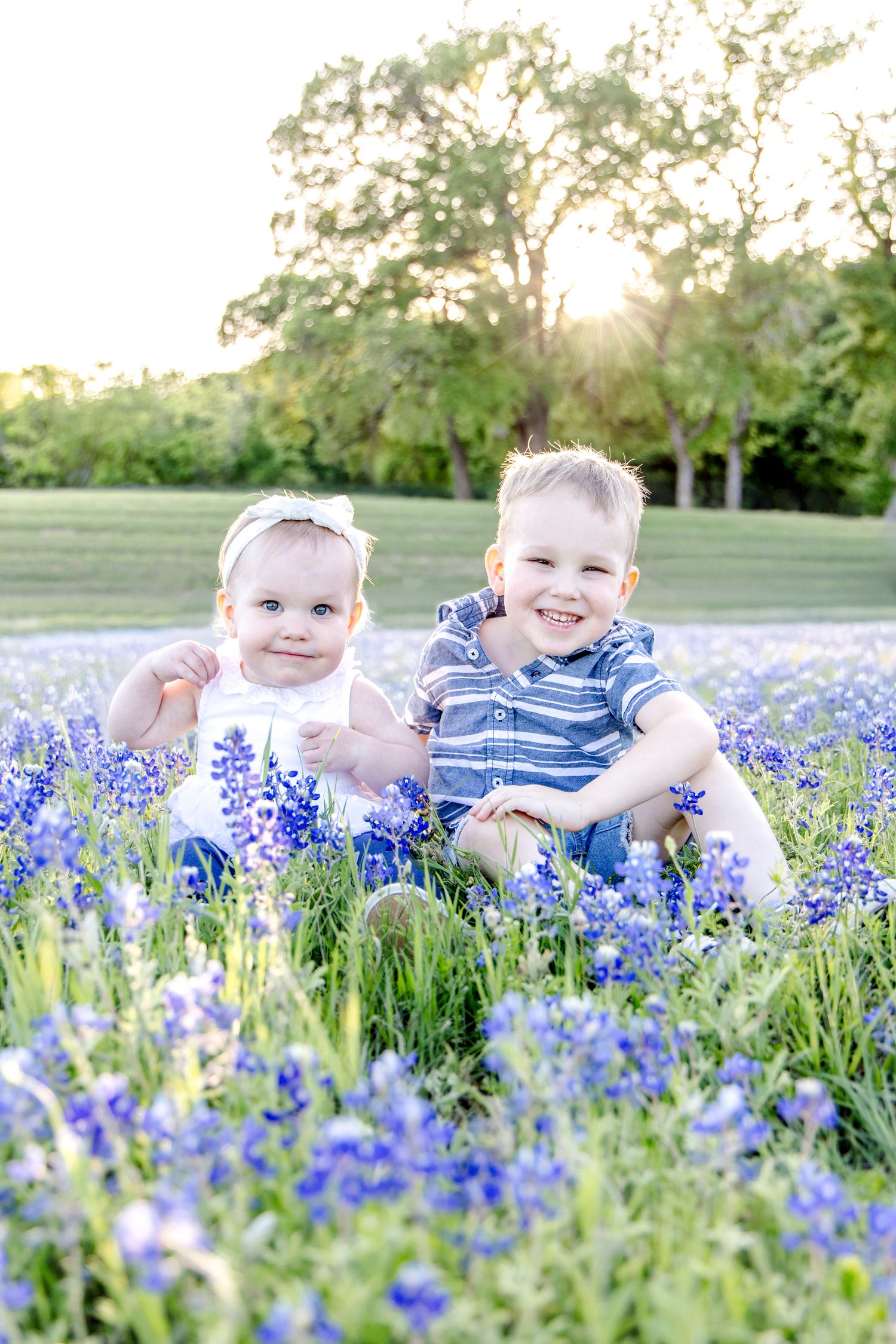 Two young children sitting together in a field of blooming purple bluebonnets during golden hour.