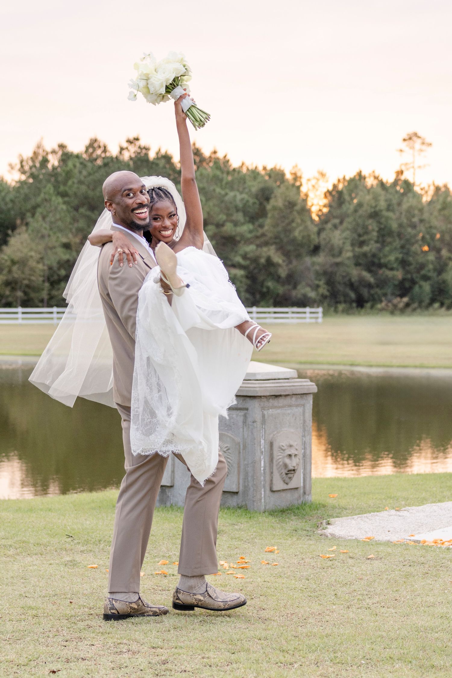 A joyful wedding couple celebrates by a pond at sunset, with the bride holding up her bouquet as she's lifted in the groom's arms.