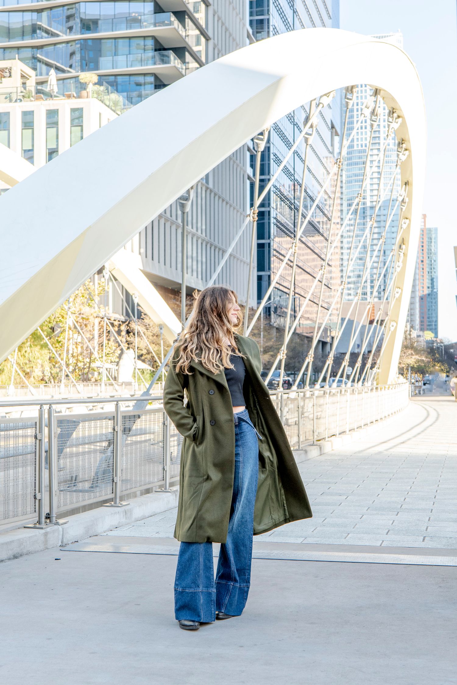 Person in long green coat poses on modern bridge with striking architectural elements.