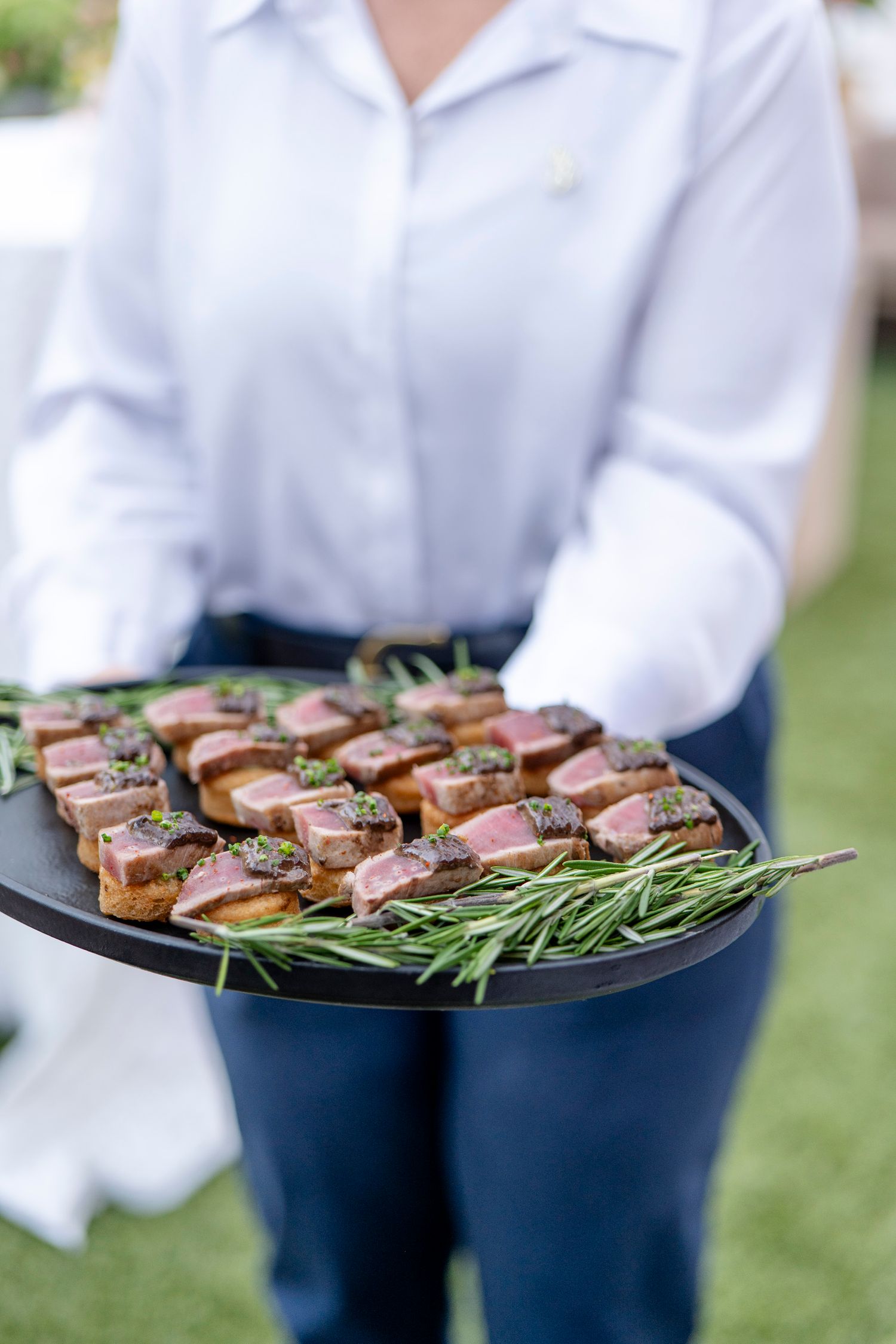 Server holding tray of sliced meat appetizers garnished with fresh rosemary sprigs.