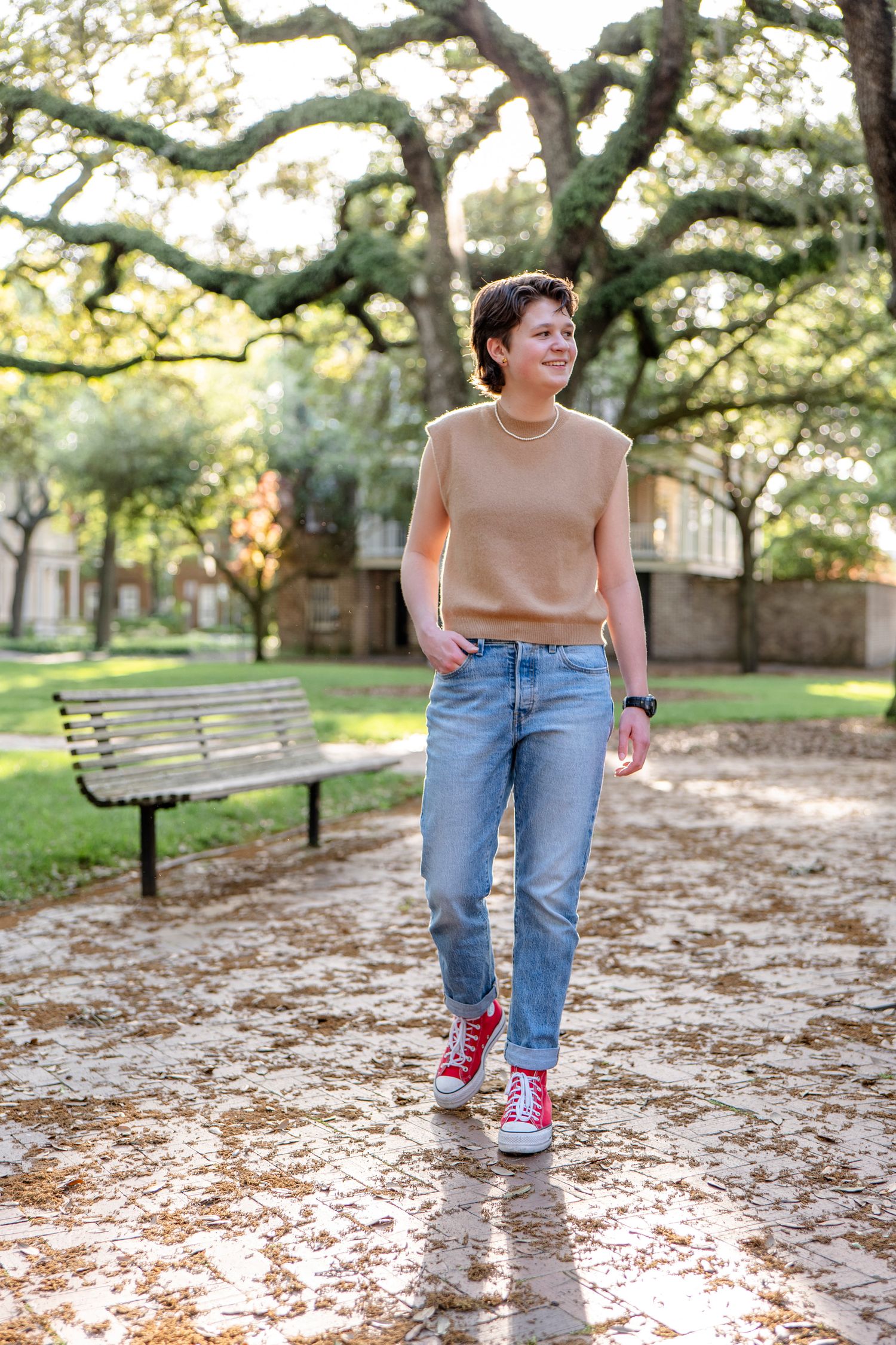 A person in casual clothing walking along a tree-lined path with a park bench.