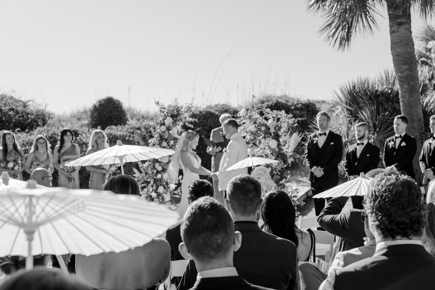 Black and white photo of guests with parasols during an elegant outdoor wedding ceremony.