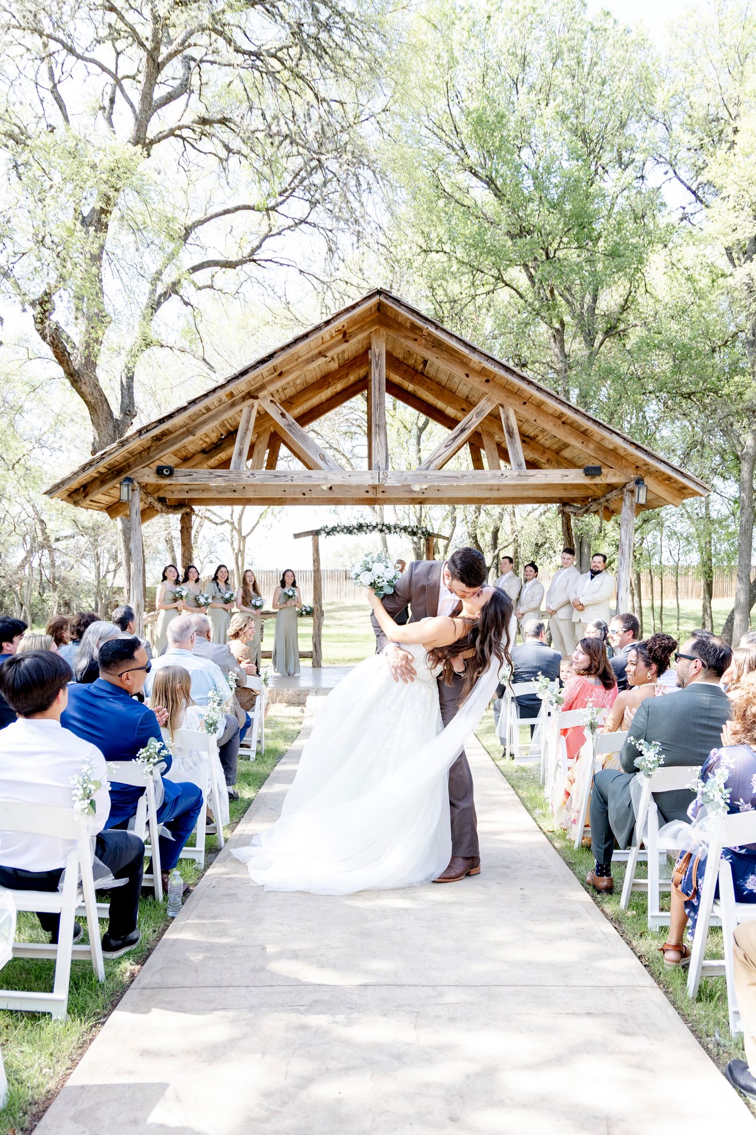 A rustic outdoor wedding ceremony takes place under a wooden pavilion surrounded by trees with white chairs for guests.