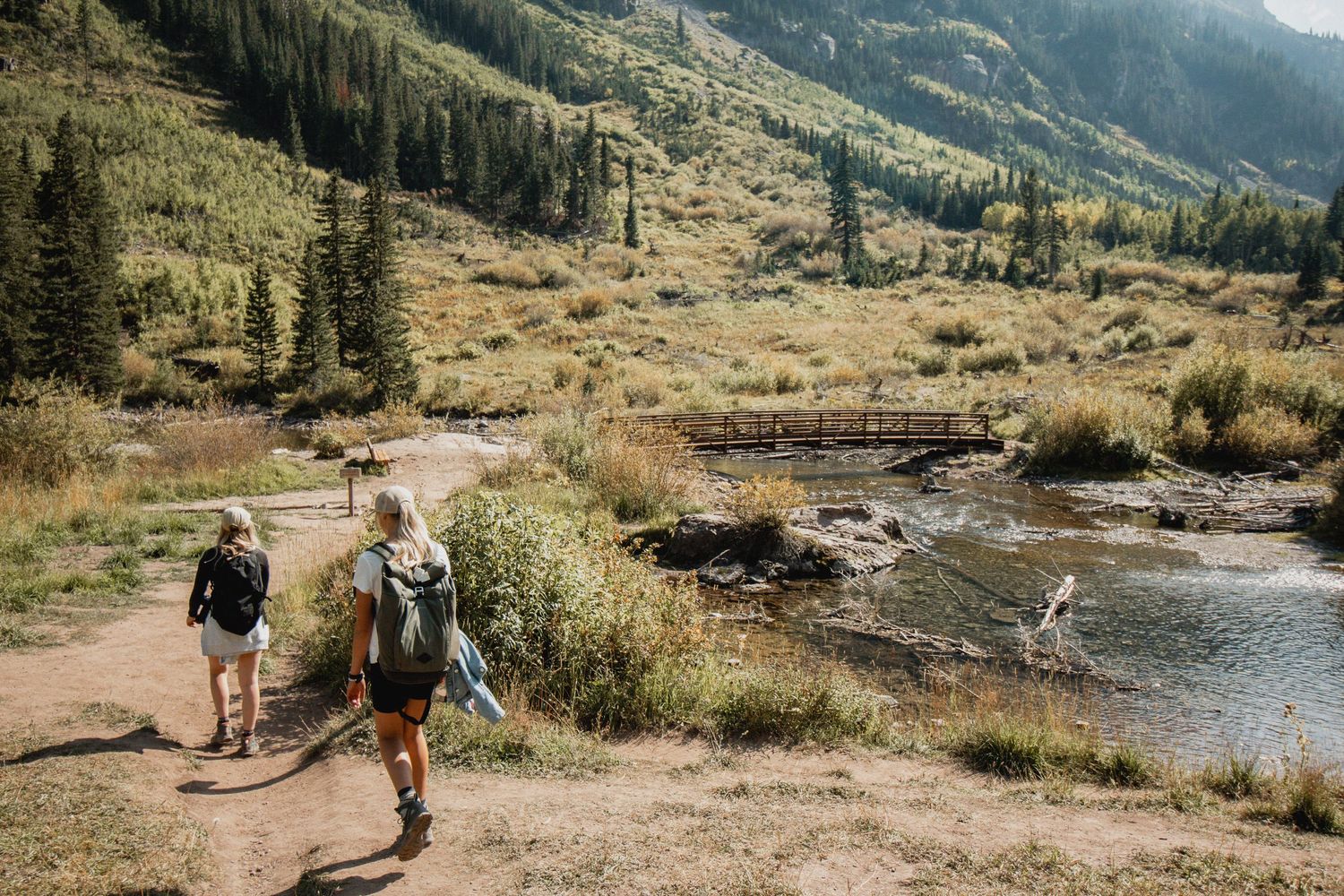 Zwei Menschen wandern an einem Fluss mit Wald und Bergen im Hintergrund.