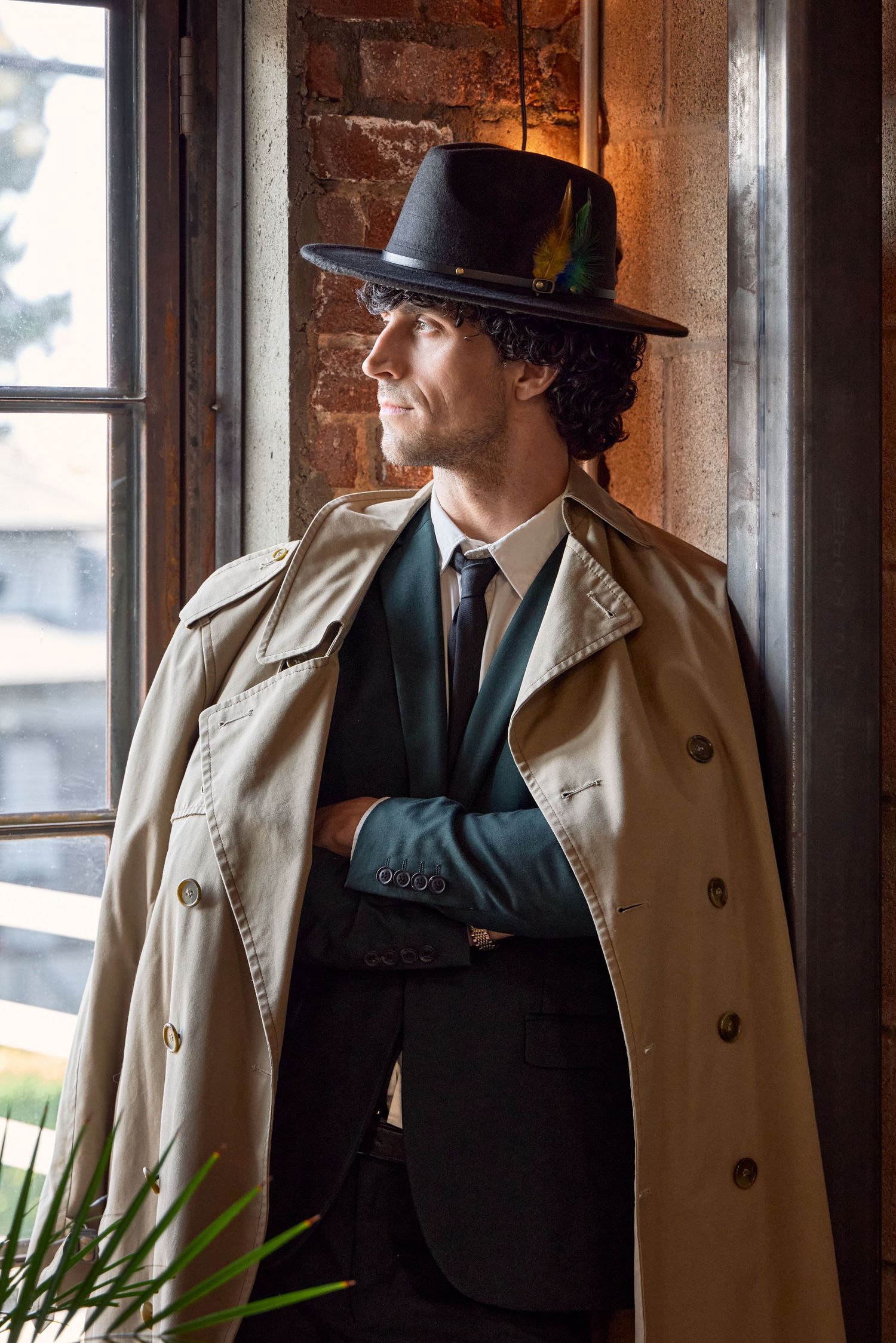 Stylish man in beige trench coat, black fedora with feather, and dark suit stands by window with arms crossed.