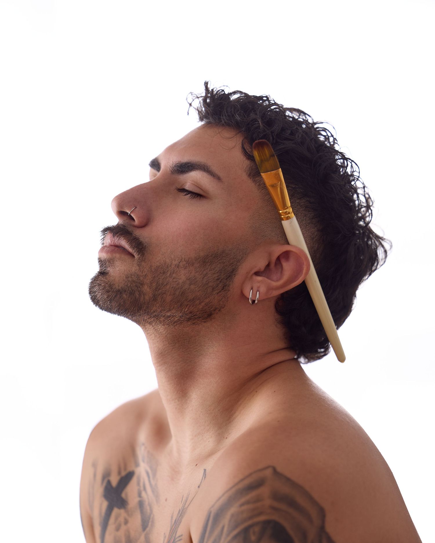 Person with curly hair and tattoos poses with closed eyes and a yellow comb in their hair against white background.