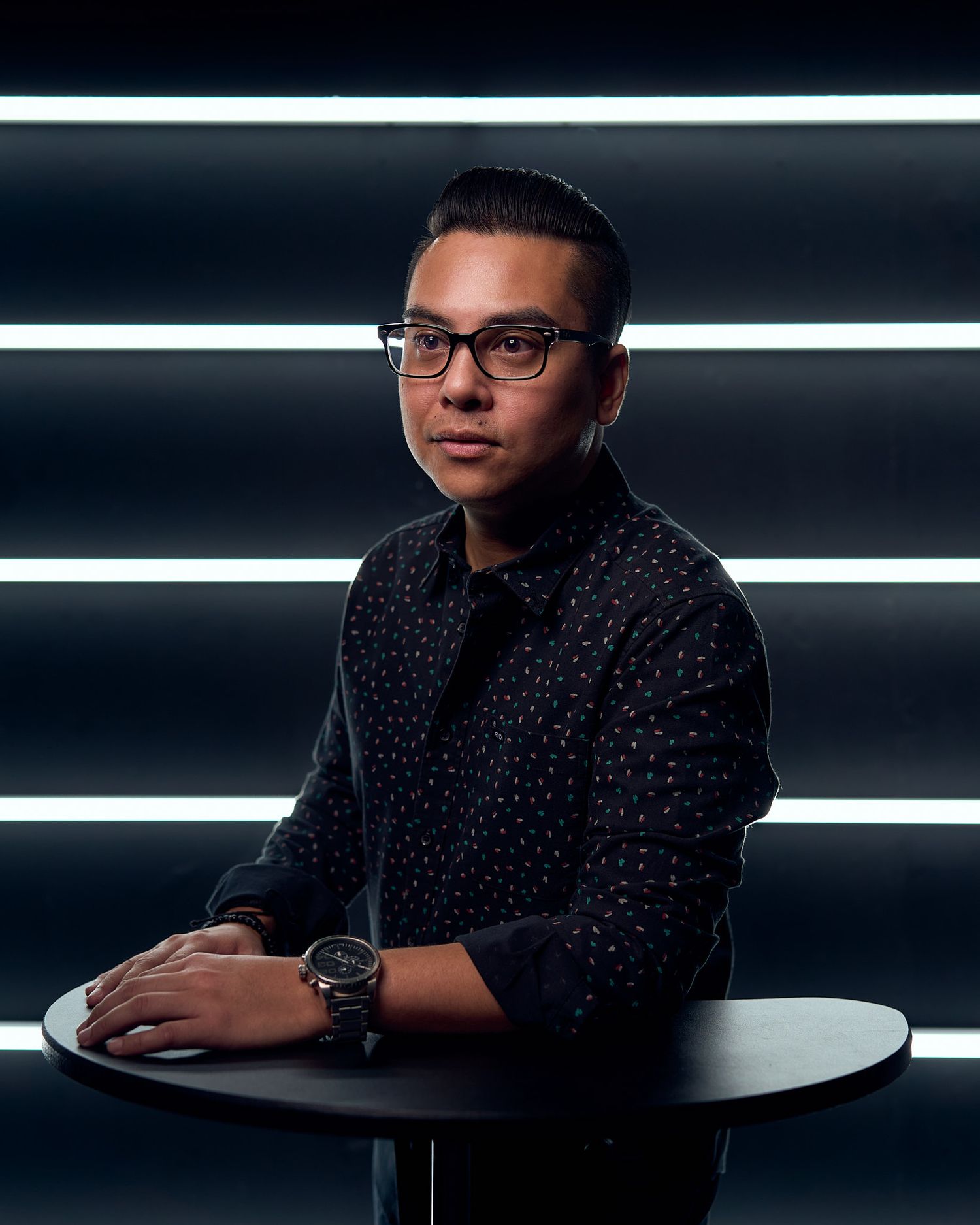 Professional seated at a modern desk with glasses and dark polka dot shirt in dramatic lighting.