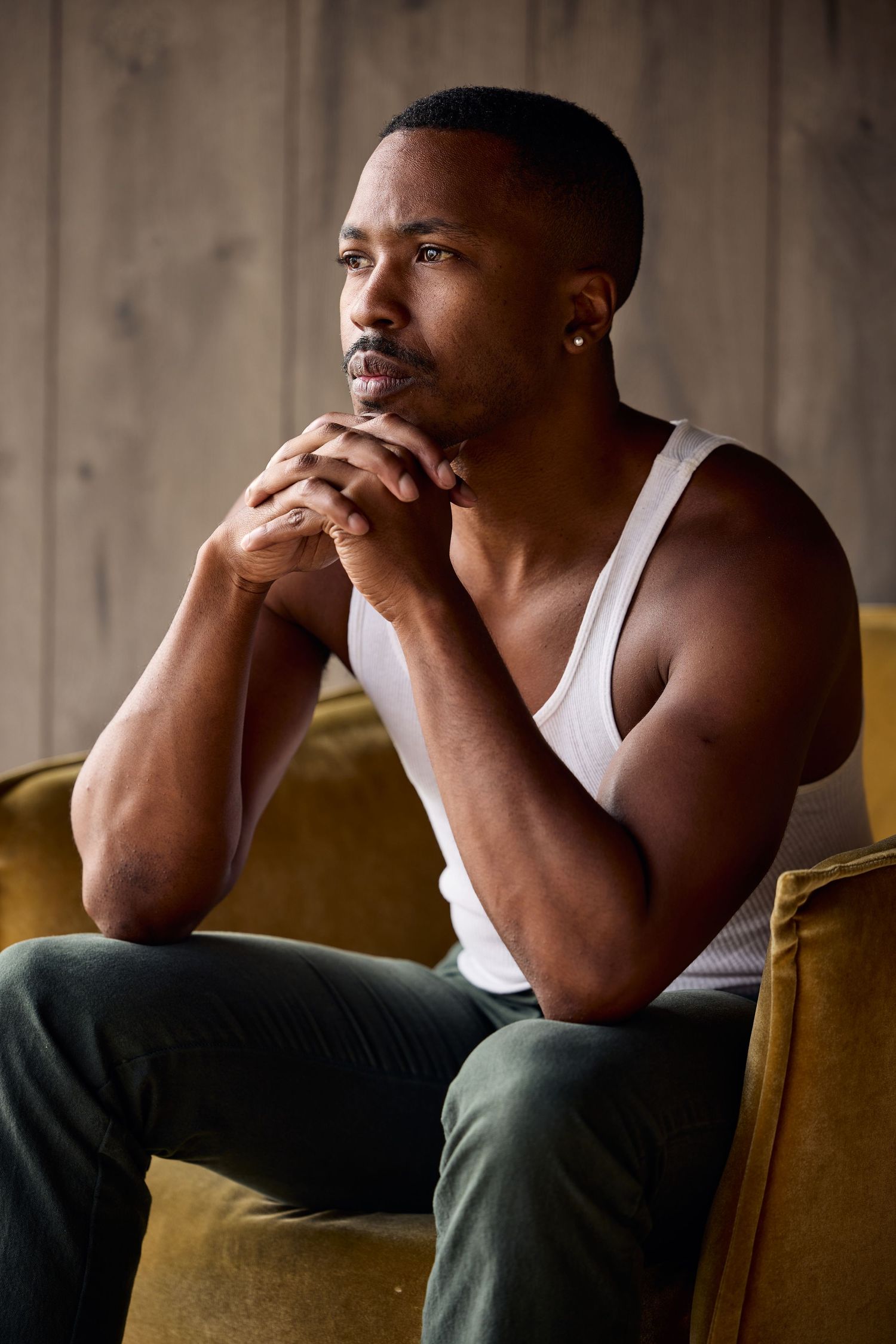 Muscular Black man in white tank top sits pensively on velvet chair, hands clasped under chin, wearing earring.