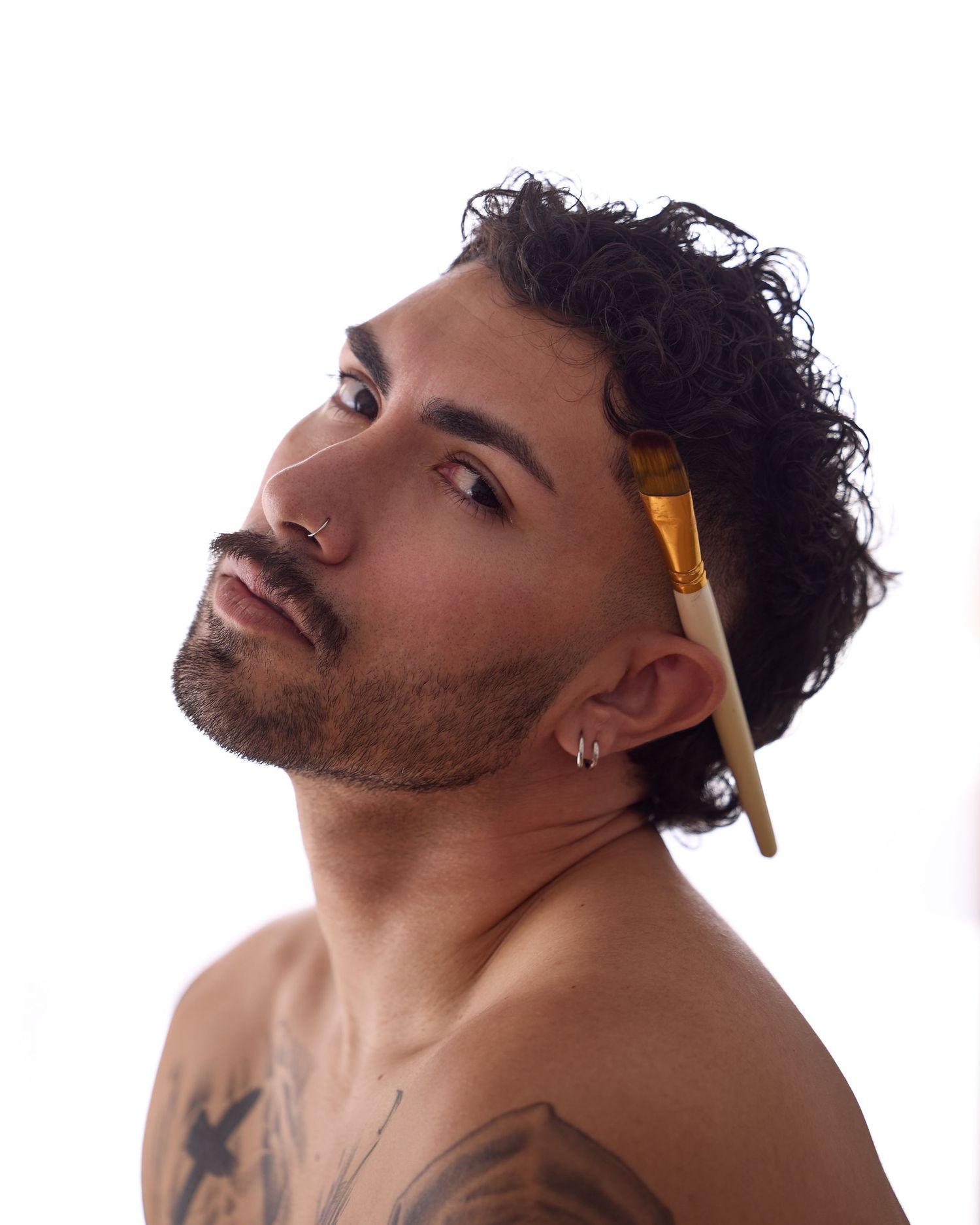 Shirtless man with curly hair, beard, ear piercing, and tattoos holds a paintbrush near his face against white background.
