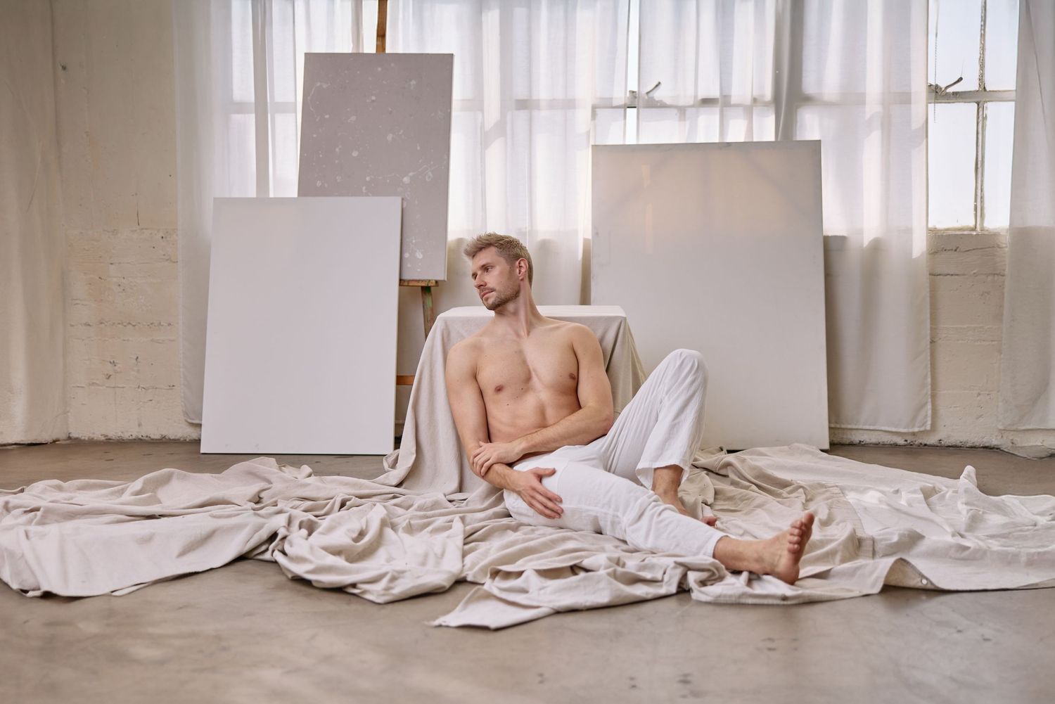 Shirtless man sitting on white draped fabric floor surrounded by blank canvases in a bright art studio.