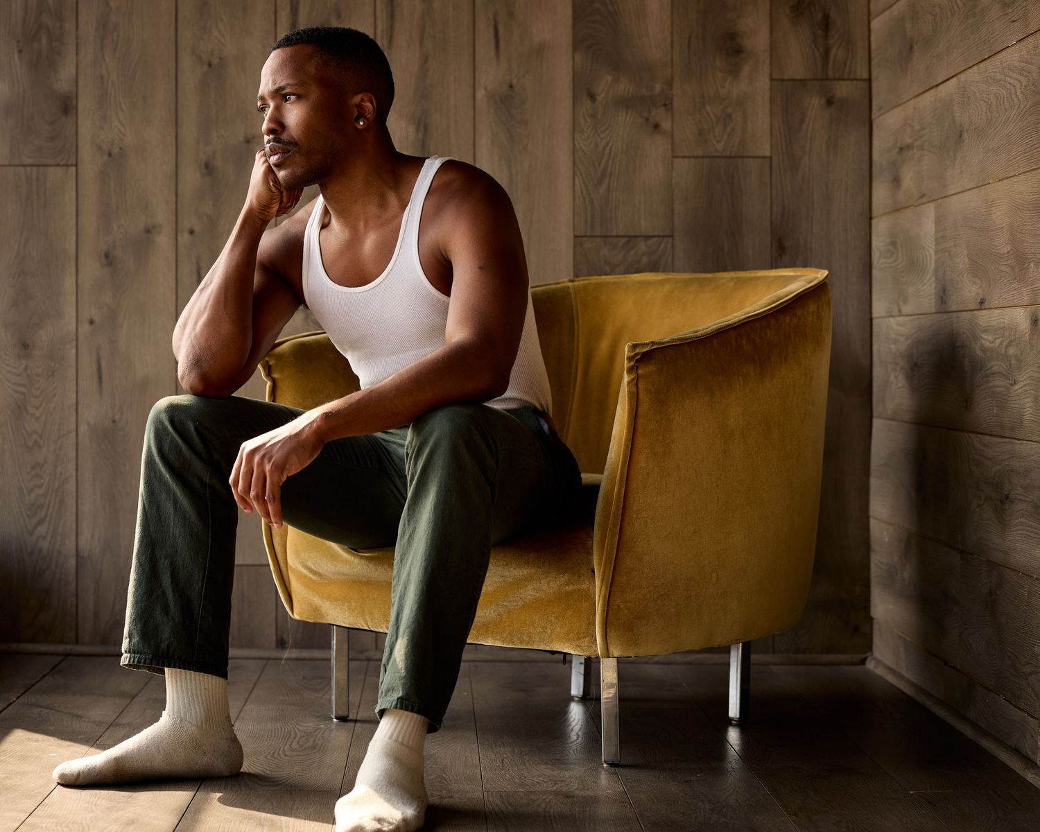Figure in white tank top seated on yellow chair against wooden wall.