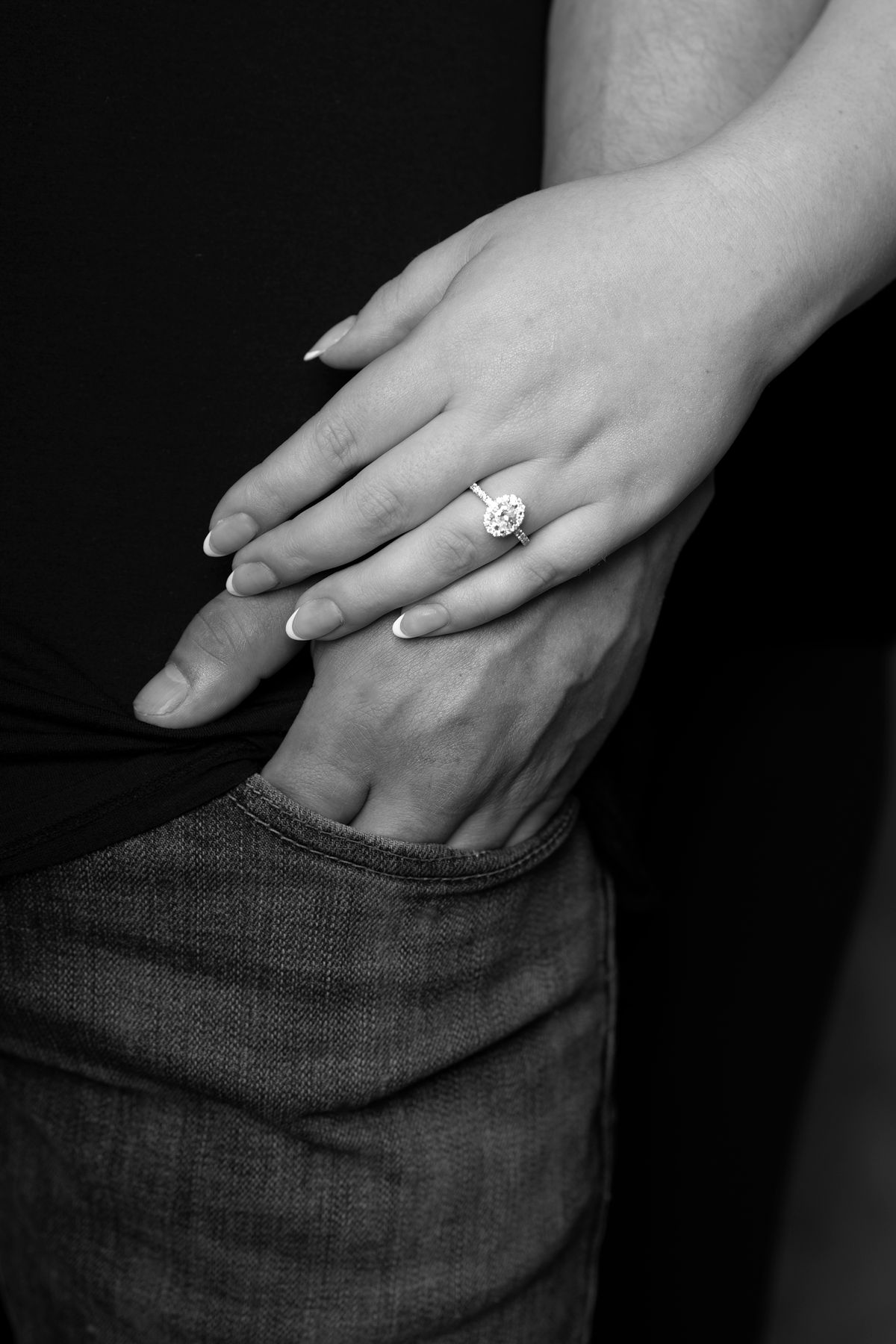 A hand wearing an engagement ring shown in ablack and white photos resting against dark fabric.