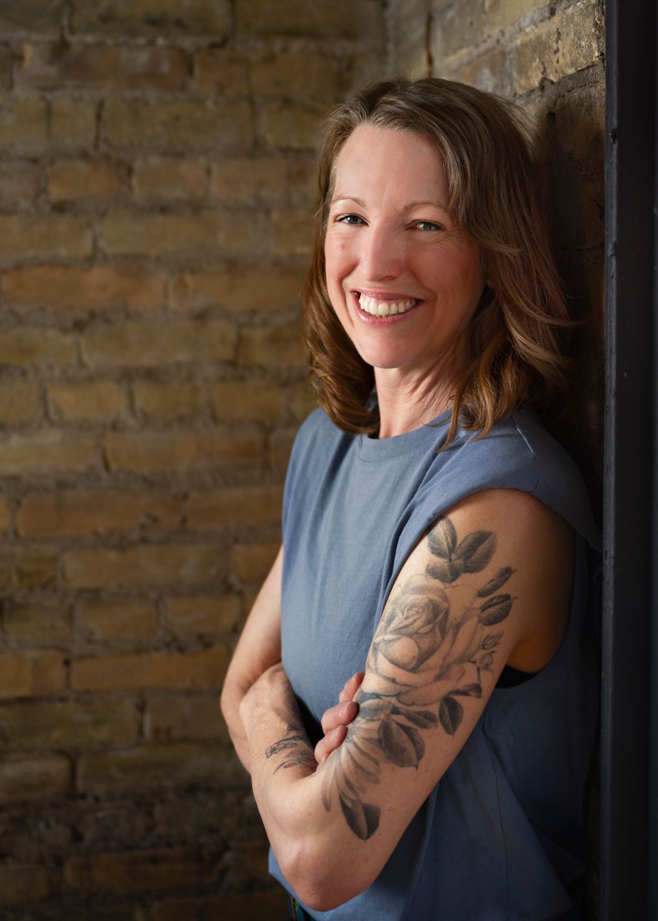 A woman in a blue sleeveless top smiles and poses against a brick wall showing off her floral arm tattoo.