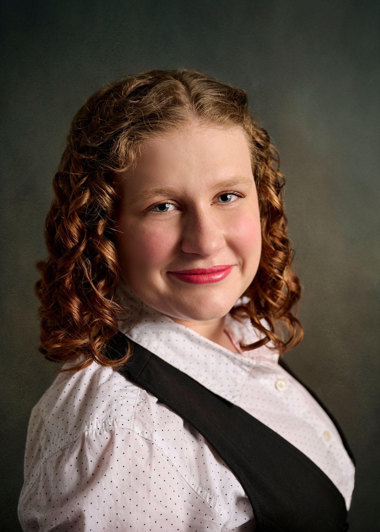 A portfolio headshot showing a person in a white top with curly hair posing against a dark background.