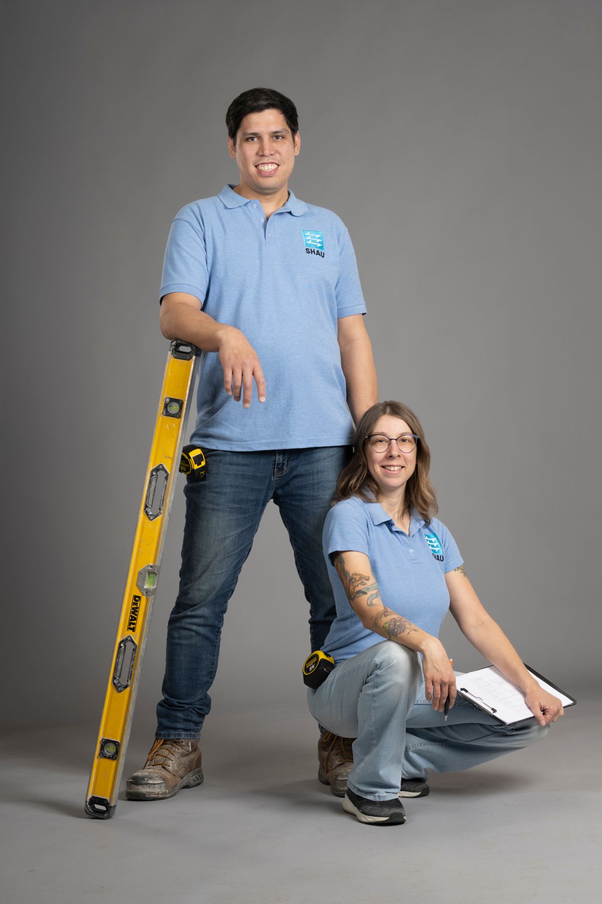 Two construction workers in blue shirts holding a yellow level and clipboard on a gray background.