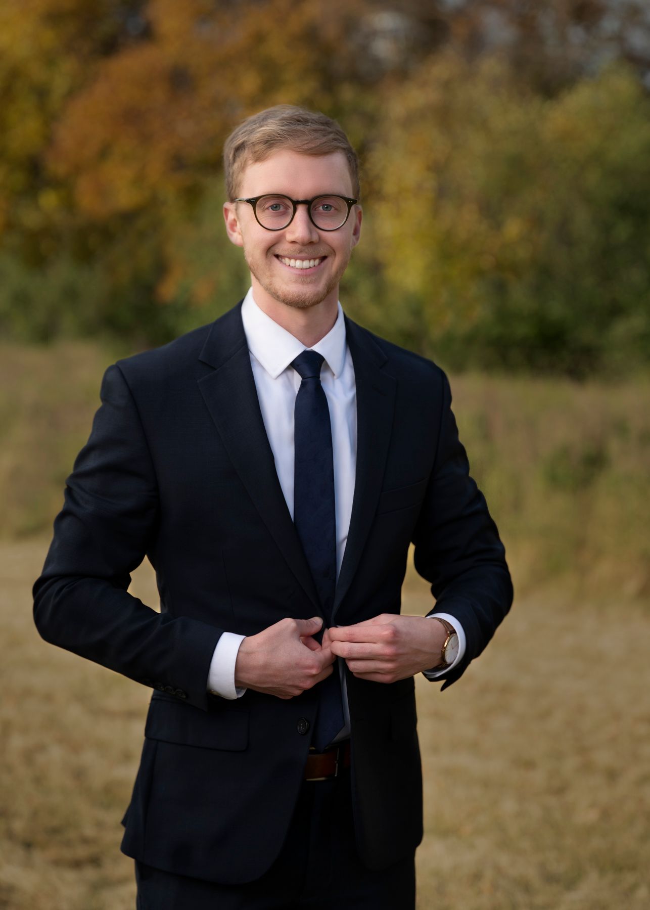Professional wearing a black suit and tie poses outdoors against an autumn background..