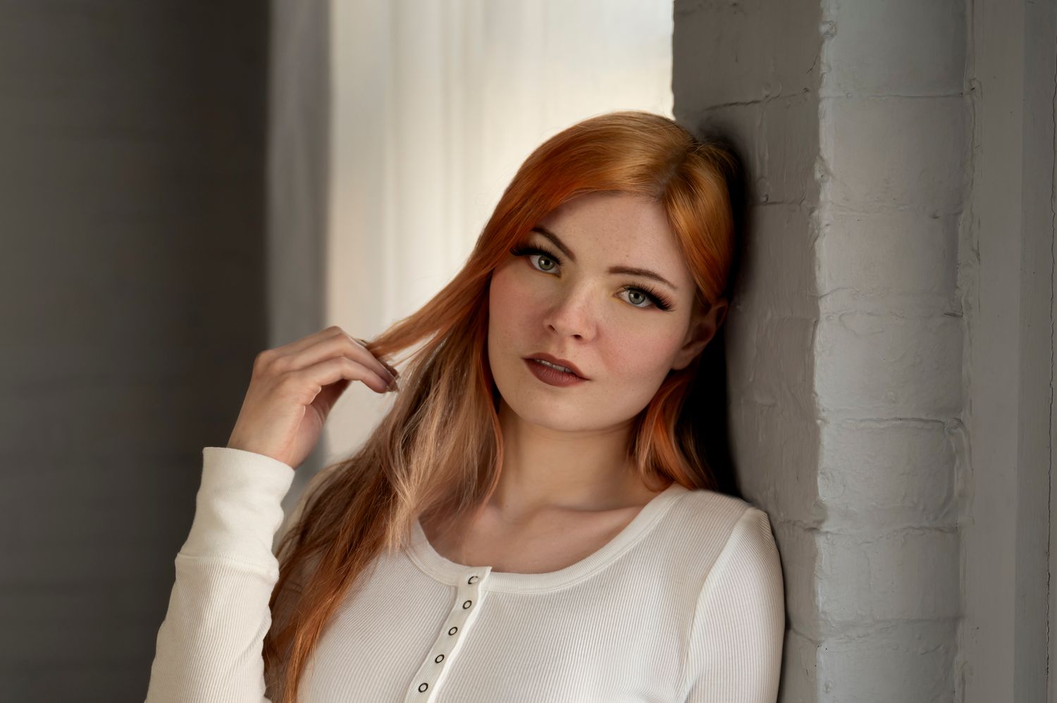 A woman with red hair in a white sweater poses against a white wall in a soft, natural light setting.