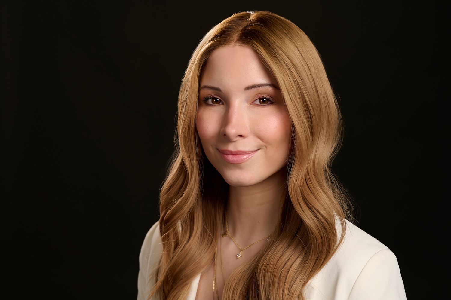 A professional headshot showing a woman with long blonde hair and a warm smile against a dark background.