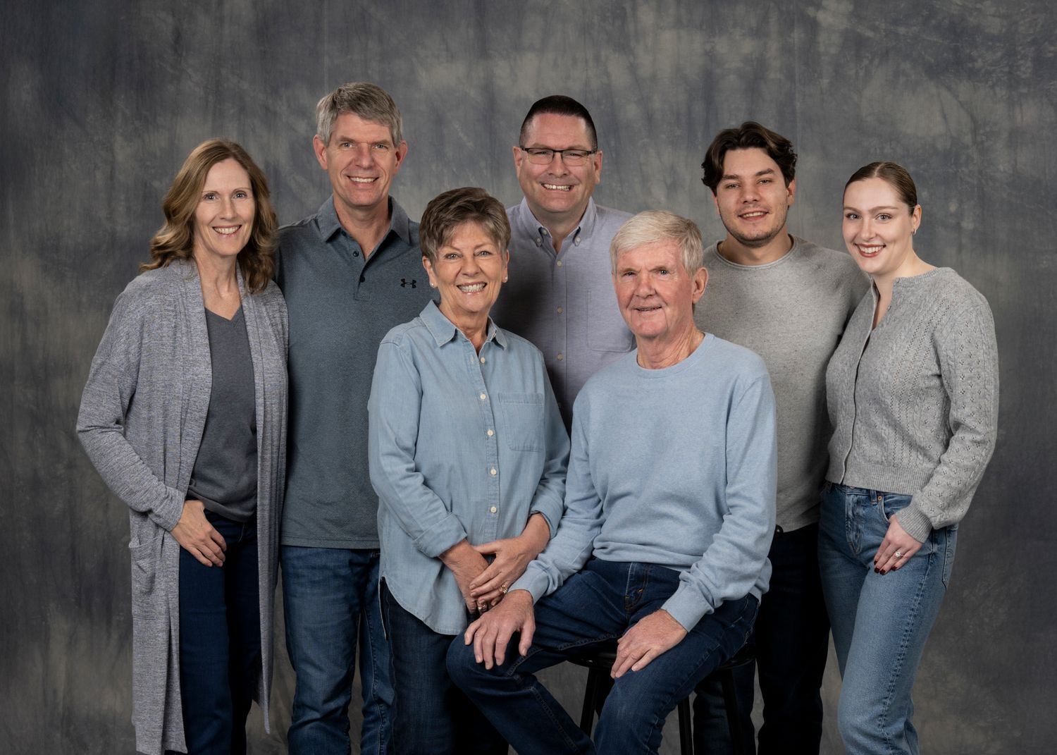 Family group wearing coordinated blue and gray outfits poses together against textured gray backdrop for portraits.