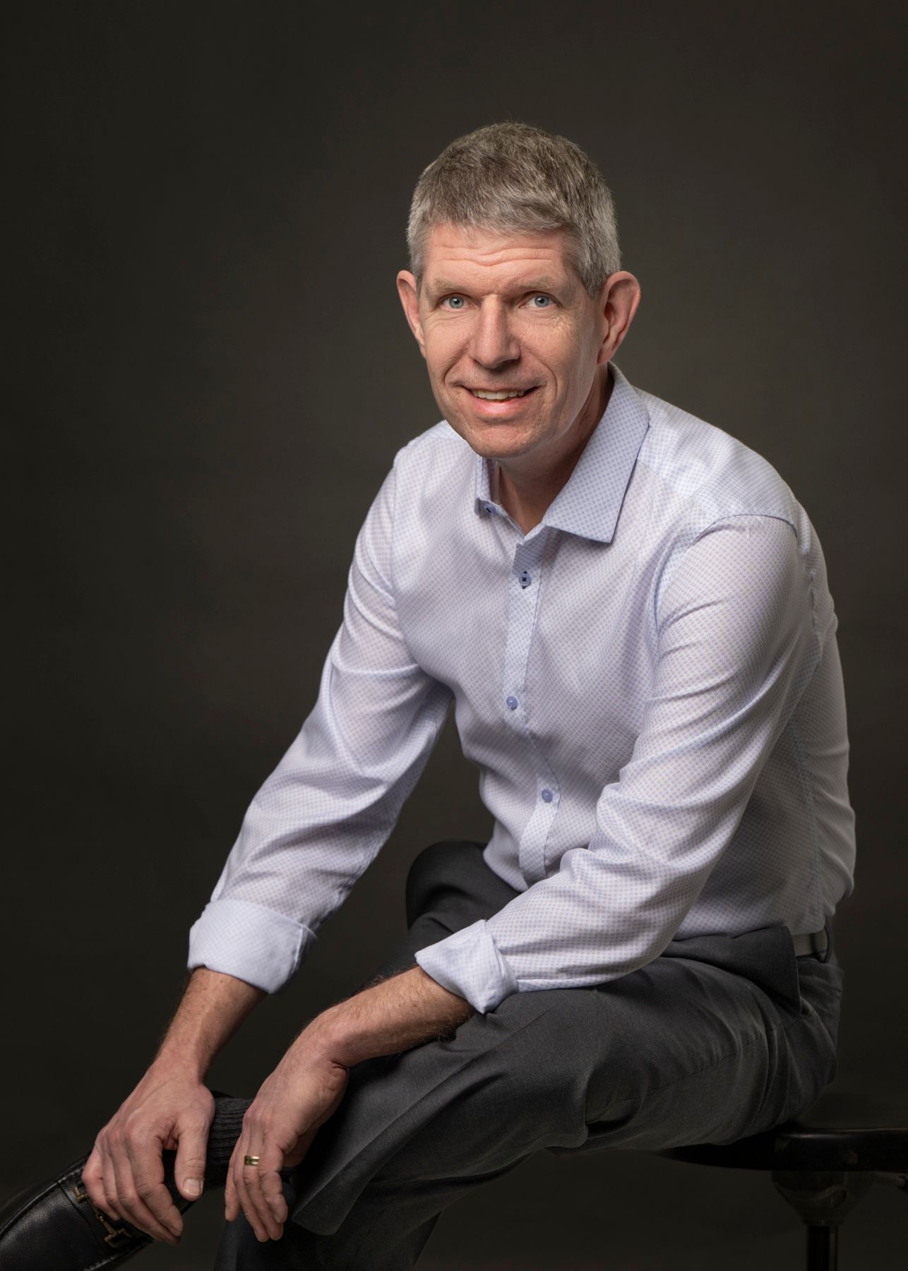 Professional studio portrait of someone in a light colored dress shirt sitting casually against dark background.