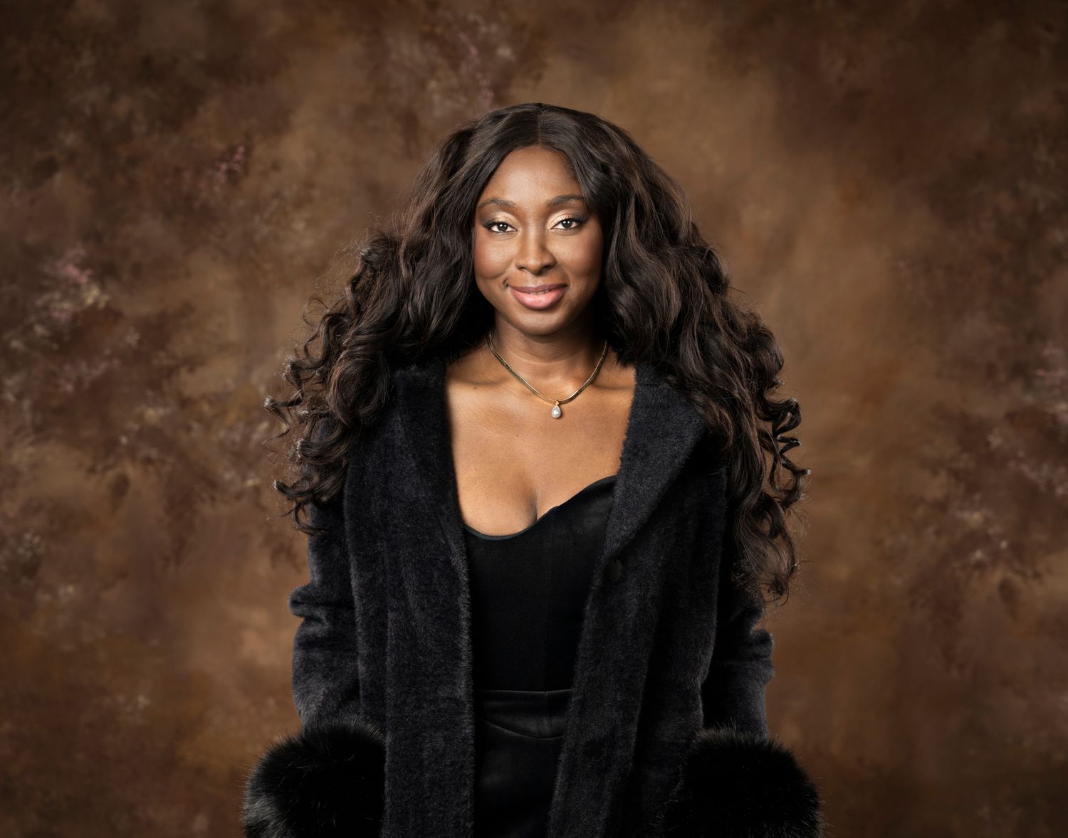 A glamorous portrait against a brown backdrop features a person in a black fur coat and velvet top with flowing dark hair.